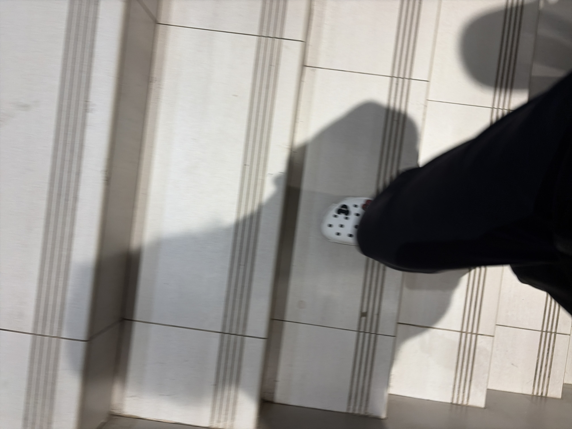 A first-person perspective shot shows a person's legs and feet descending a tiled staircase. The individual is wearing black pants and white clogs with black polka dots. The staircase tiles are light beige with horizontal grey lines etched into them. Shadows from the person and an unseen light source are cast on the stairs. The scene is not identifiable as Moscow, Russia.