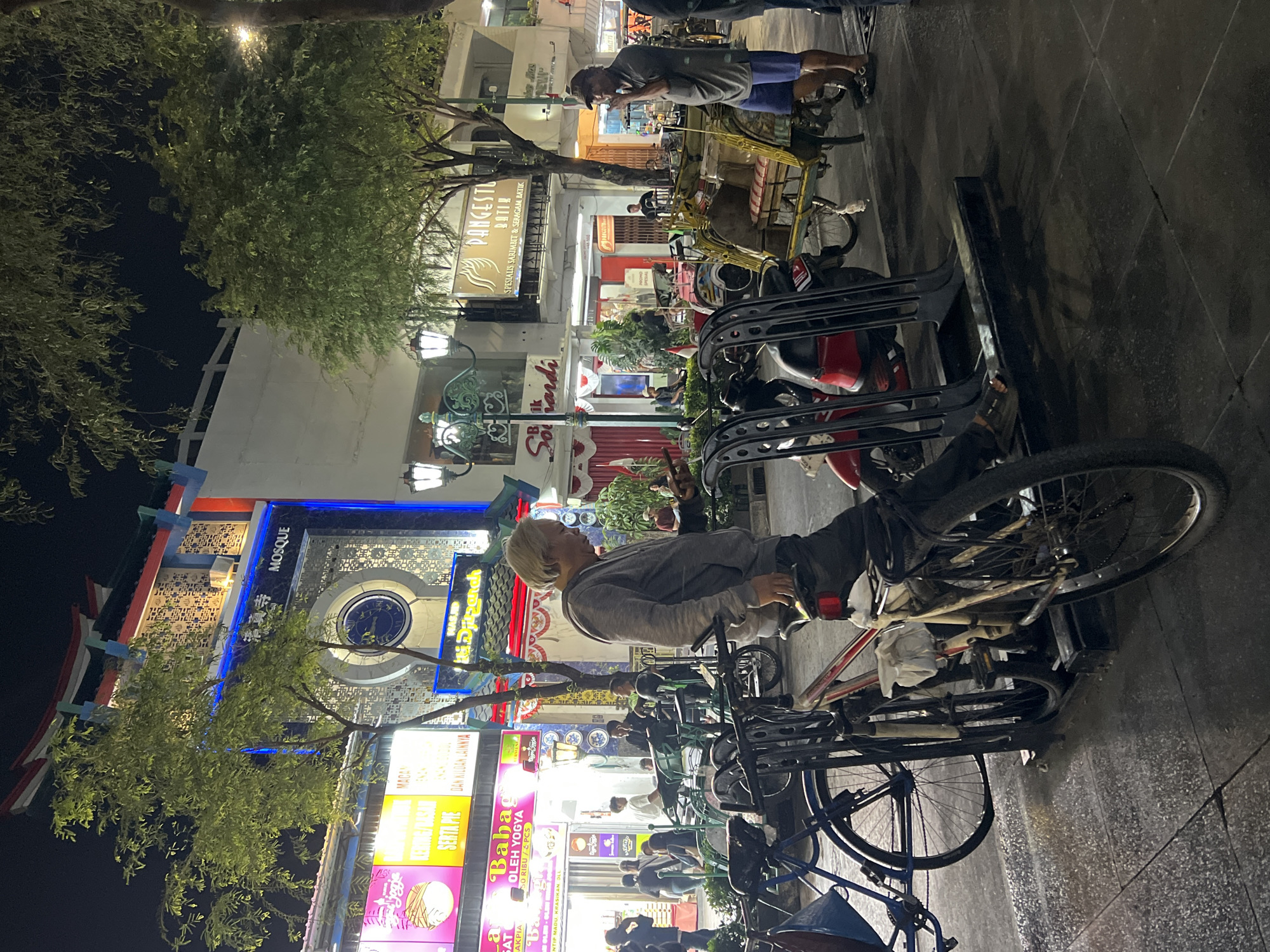 This image depicts a nighttime scene in a pedestrian area of Yogyakarta, Indonesia, likely a busy commercial or entertainment district. The foreground shows a bicycle rack filled with several bicycles and a motorcycle, indicating a place where people park their vehicles. A man with graying hair is standing next to a bicycle, looking down. To his right, another man in a cap and blue shorts is standing near a pedicab, possibly a driver waiting for passengers or taking a break.
The background reveals a lively street with various shops and businesses illuminated by bright signage. One prominent building features a distinctive architectural style with a curved roof, a large circular motif, and signage that includes the word "MOSQUE," suggesting a religious or cultural landmark. Other shops display colorful advertisements, with some text visible in Indonesian, such as "Baba" and "Oleh Yogya." The overall atmosphere suggests a vibrant urban environment after dark, with people out and about. The wet pavement indicates recent rain or cleaning, adding a reflective quality to the scene.