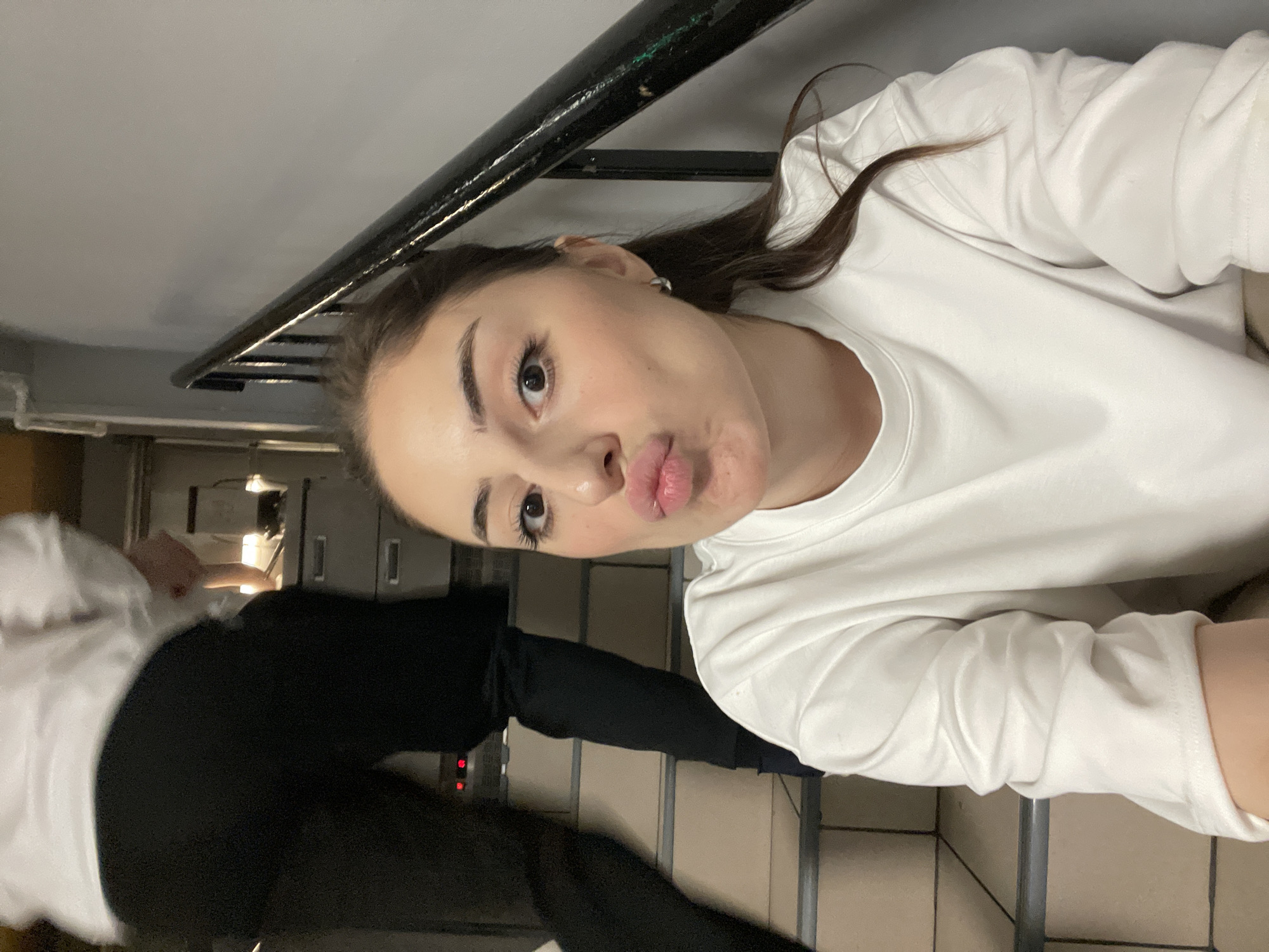 A young woman with dark hair and light skin is taking a selfie. She is wearing a white long-sleeved shirt. She is making a kissing face. The background appears to be a stairwell with a black handrail and beige stairs. Another person, wearing a white shirt and black pants, is partially visible in the lower left corner of the image. The location is Moscow, Russia.