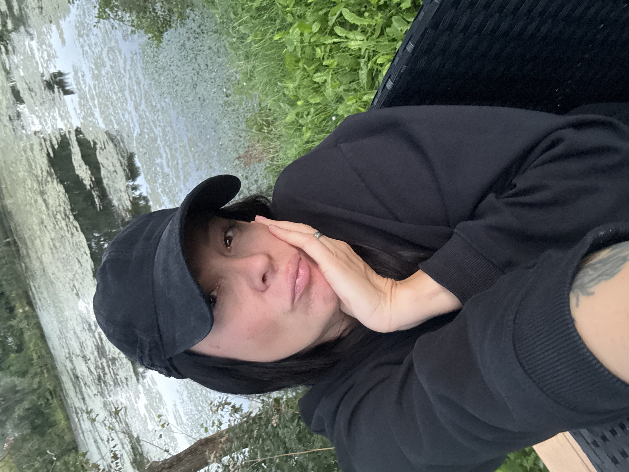 A woman wearing a black baseball cap and a black sweatshirt is seated outdoors, with her right hand resting on her cheek. She is looking towards the camera with a pout. The background features a body of water covered in green algae, with trees and vegetation on the banks. The scene appears to be a park or a natural area. The specific location in Chaikovsky, Russia, is not discernible from the image.