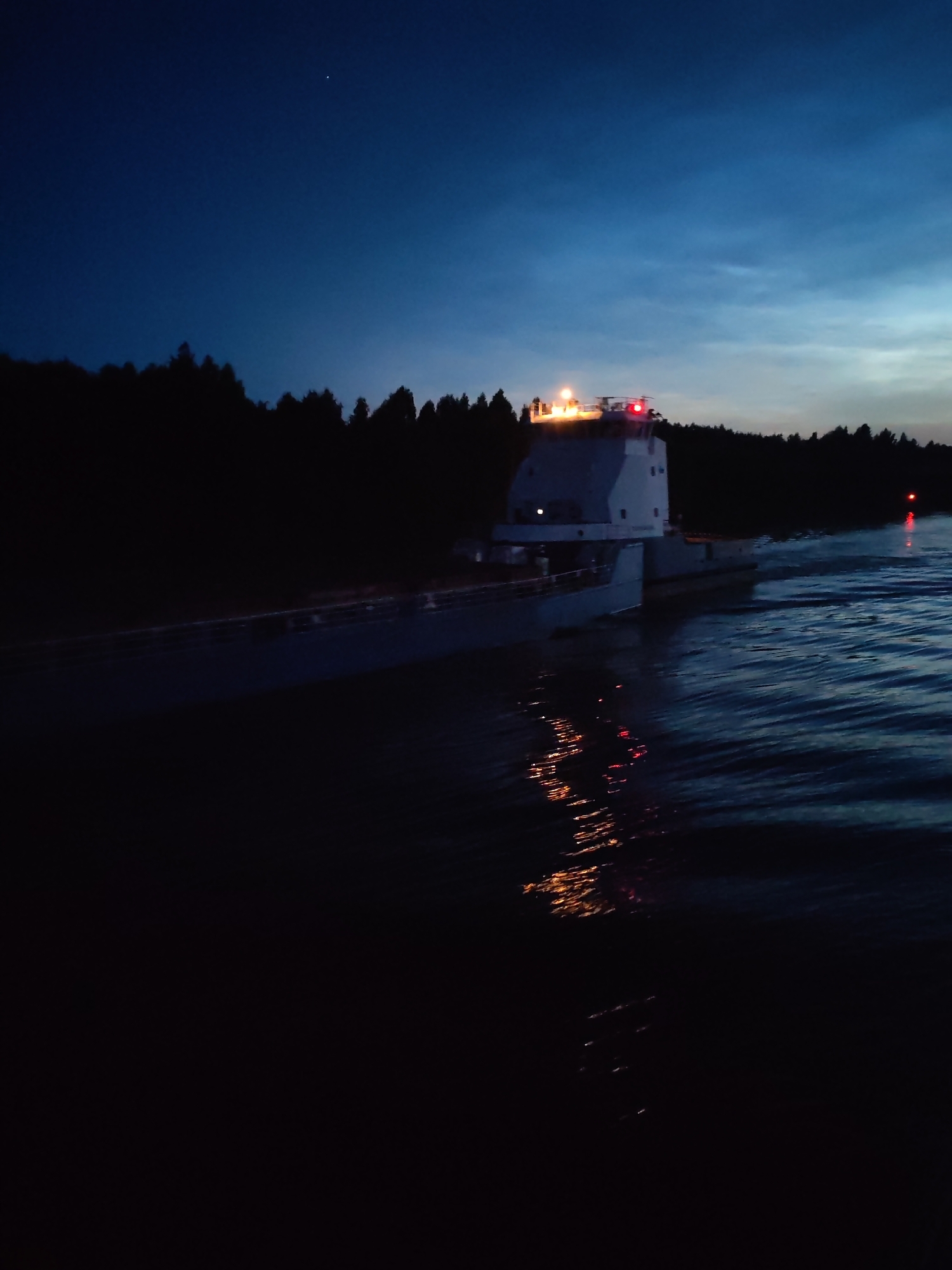 At dusk, a long, white vessel navigates on the dark, rippling waters of the Vytegra River, near the city of Vytegra, Russia. The ship, with a prominent white superstructure topped with illuminated lights, is silhouetted against a deep blue sky and the dark, tree-lined shore. The water reflects the ship's lights and the faint glow of the sky, creating streaks of orange and white on the surface. A single red navigation buoy is visible in the distance, marking the waterway. The overall scene suggests a calm evening on the river, with the vessel proceeding on its journey.