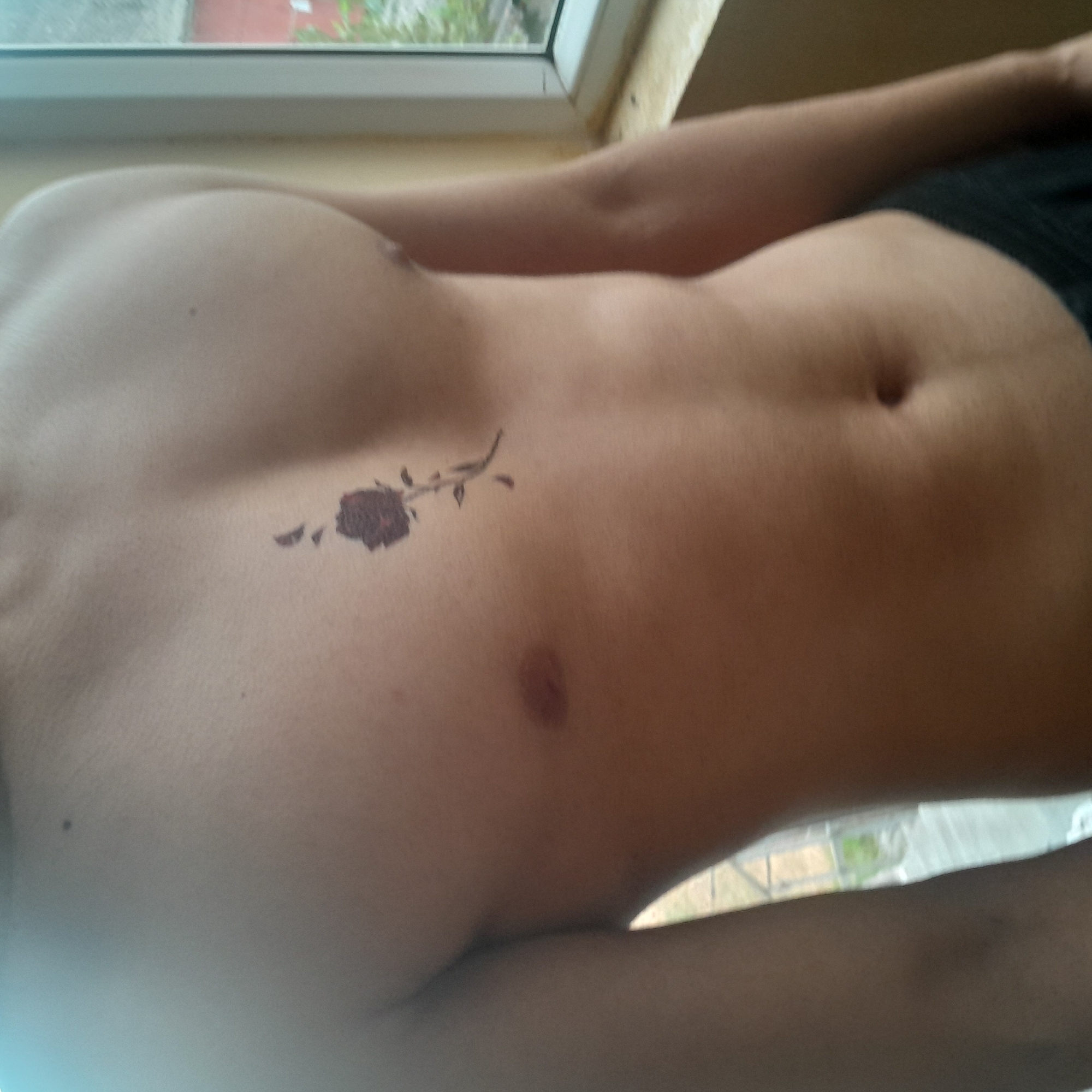 A close-up, medium shot captures the bare chest and upper abdomen of a person, with a small, dark rose tattoo on the sternum. The tattoo is positioned vertically, with the stem extending downwards and the flower head at the top. The person's skin is smooth and tanned, and their abdominal muscles are subtly defined. To the right of the frame, a portion of a window with a light-colored frame is visible, and through the glass, some greenery and hints of buildings suggest an outdoor environment. The lighting is soft, coming from the right side, casting subtle shadows across the skin. The scene is devoid of any context regarding location within Astara, Azerbaijan, as no identifiable landmarks or specific environmental cues are present.