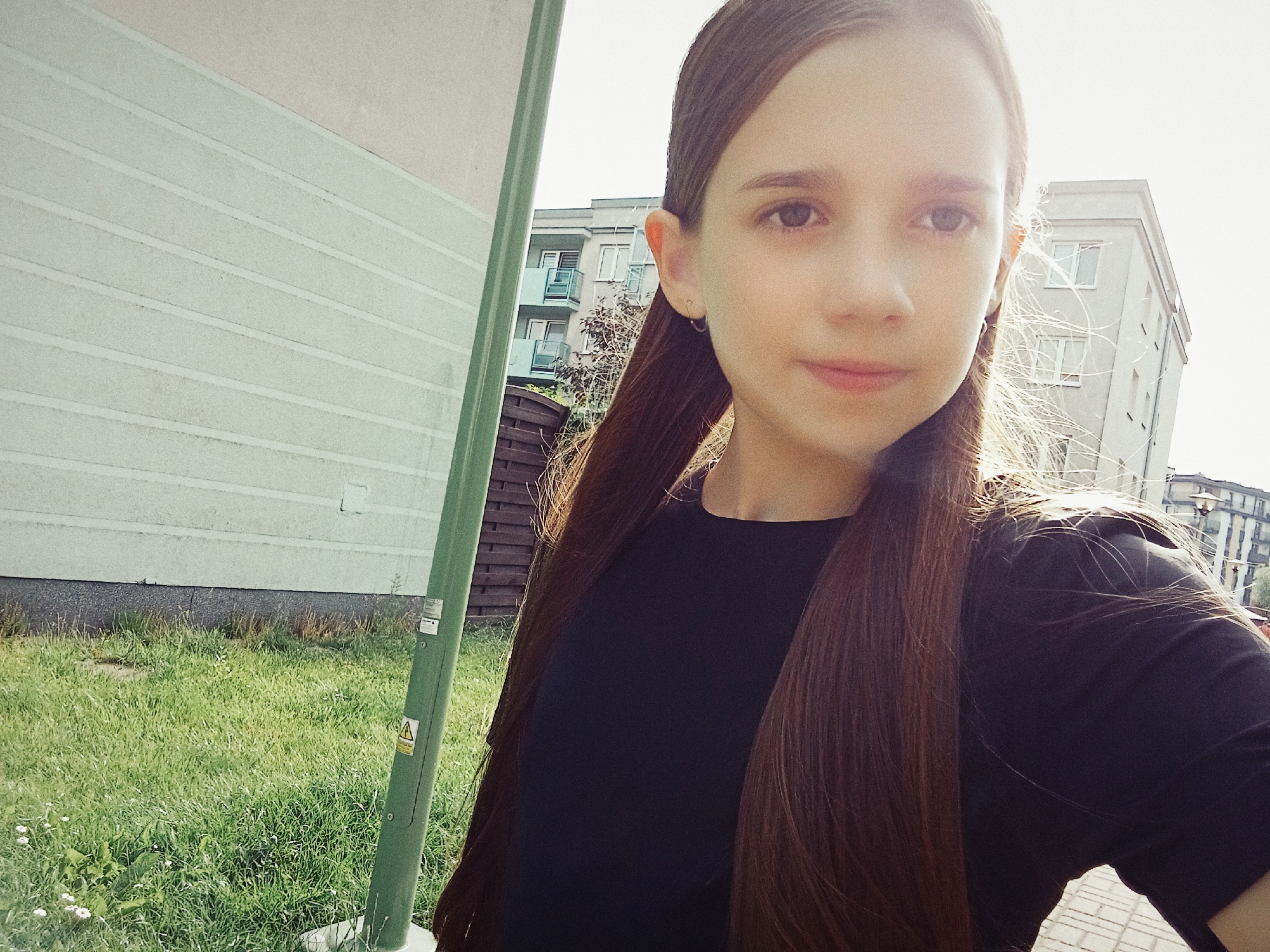 In the city of Dybów Kolonia, Poland, a young girl with long brown hair is captured in a selfie. She is wearing a dark t-shirt and her hair is styled with two low ponytails, with strands of hair catching the sunlight. Behind her, a pale green building with horizontal paneling is visible, and further in the background, modern apartment buildings with balconies rise against a bright sky. The ground is covered in green grass, and a tall green pole, possibly a street light or utility pole, stands to the left of the frame. The scene appears to be a bright, sunny day, possibly in a residential area.