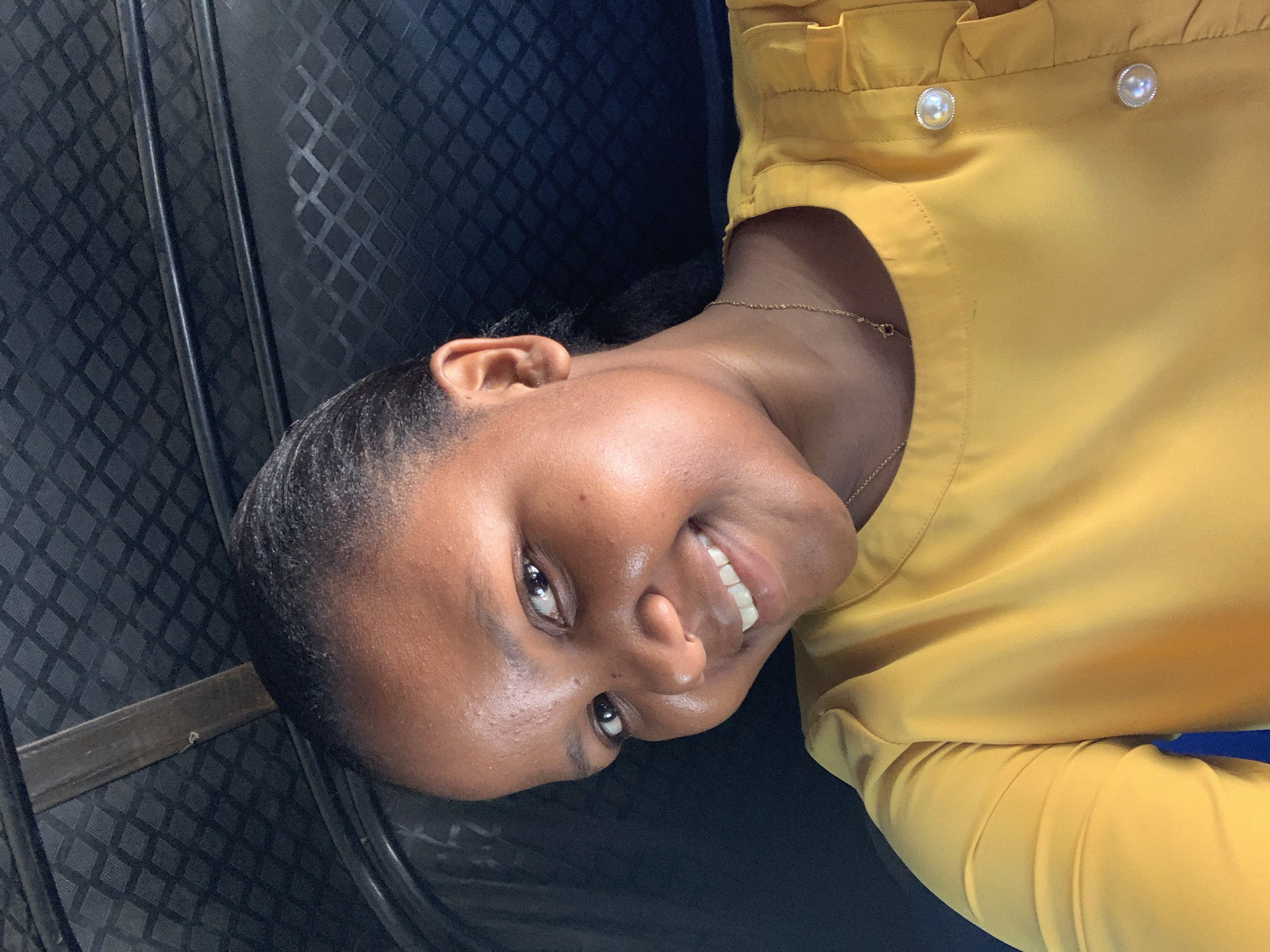 A young woman is posing for a photograph inside a vehicle. She is wearing a yellow blouse with pearlescent buttons at the collar and a delicate gold necklace. The woman has dark hair pulled back from her forehead, and she is smiling slightly, revealing her upper teeth. The background is a dark patterned surface, likely the interior of a seat or wall of the vehicle. The specific location in Dar es-Salaam, Tanzania cannot be determined from the image.