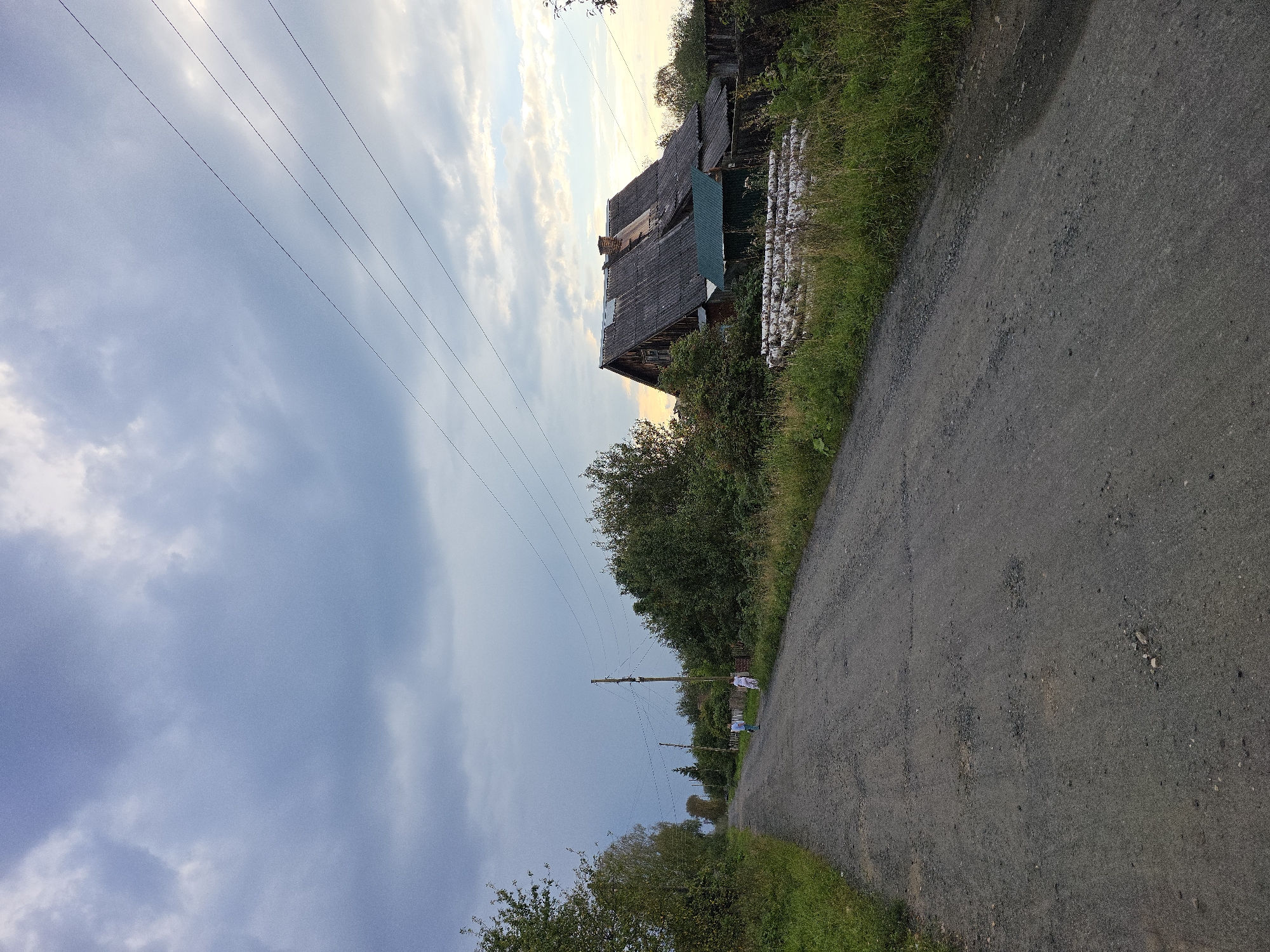 The image depicts a rural road leading towards a dilapidated wooden house in Magnitka, Russia. The road is unpaved and appears to be in poor condition, with visible potholes and loose gravel. On the right side of the road, a two-story wooden house with a gray, weathered roof is situated amidst overgrown vegetation. A pile of logs is stacked in front of the house. Electrical wires are strung overhead, supported by wooden poles. Two individuals are visible walking away from the viewer on the road. The sky is overcast with heavy clouds, suggesting impending rain or a gloomy atmosphere. The surrounding environment is characterized by dense trees and shrubs, indicating a natural or semi-natural setting.