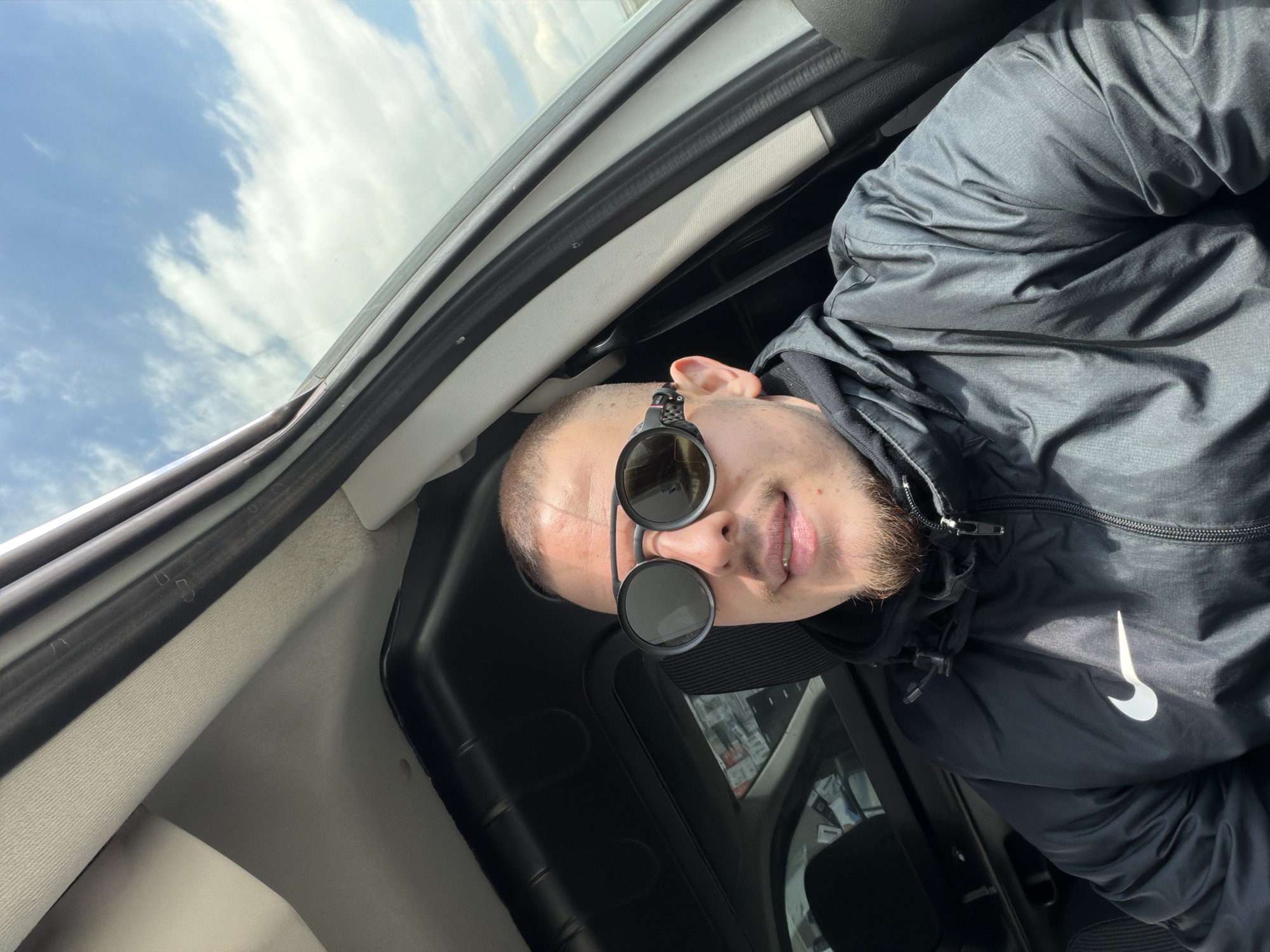 A man with short hair, a beard, and round sunglasses is inside a vehicle. He is wearing a black hooded jacket with a white Nike logo. The interior of the vehicle is visible, along with a view of the sky with clouds through the window. The location is Parvenets, Bulgaria, though this information is not discernible from the image.