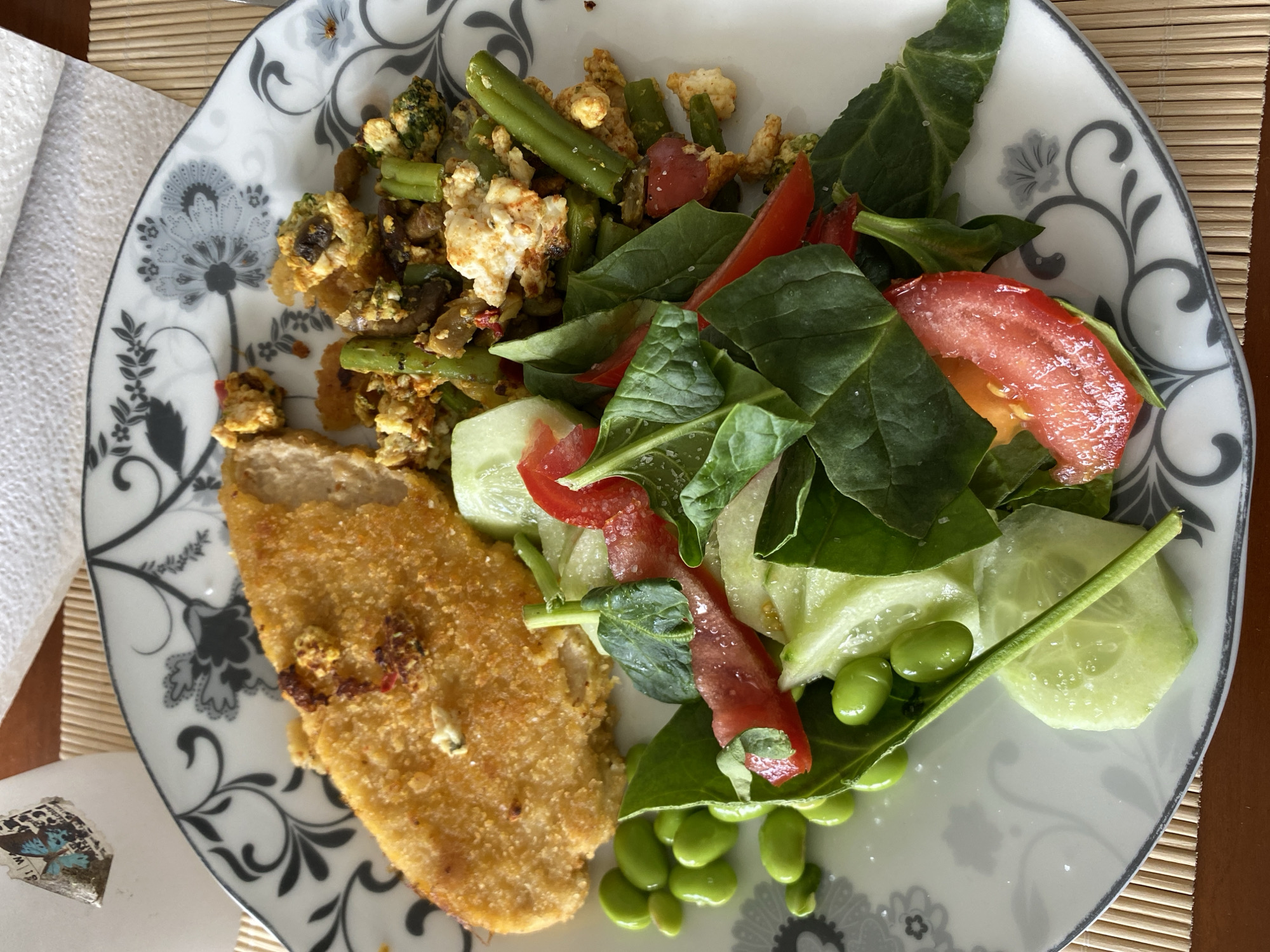 The image shows a plate of food. The plate is decorated with a floral pattern in gray and white. The food consists of a breaded cutlet, a mixture of scrambled tofu with green beans and broccoli, and a salad of spinach, tomato, cucumber, and edamame. The plate sits on a bamboo placemat. A white napkin is partially visible in the upper left corner, next to a small, decorative object, possibly a button. The setting appears to be a private residence, likely a dining table. There is no information in the image that suggests the location is in Gijón/Xixón, Spain.
