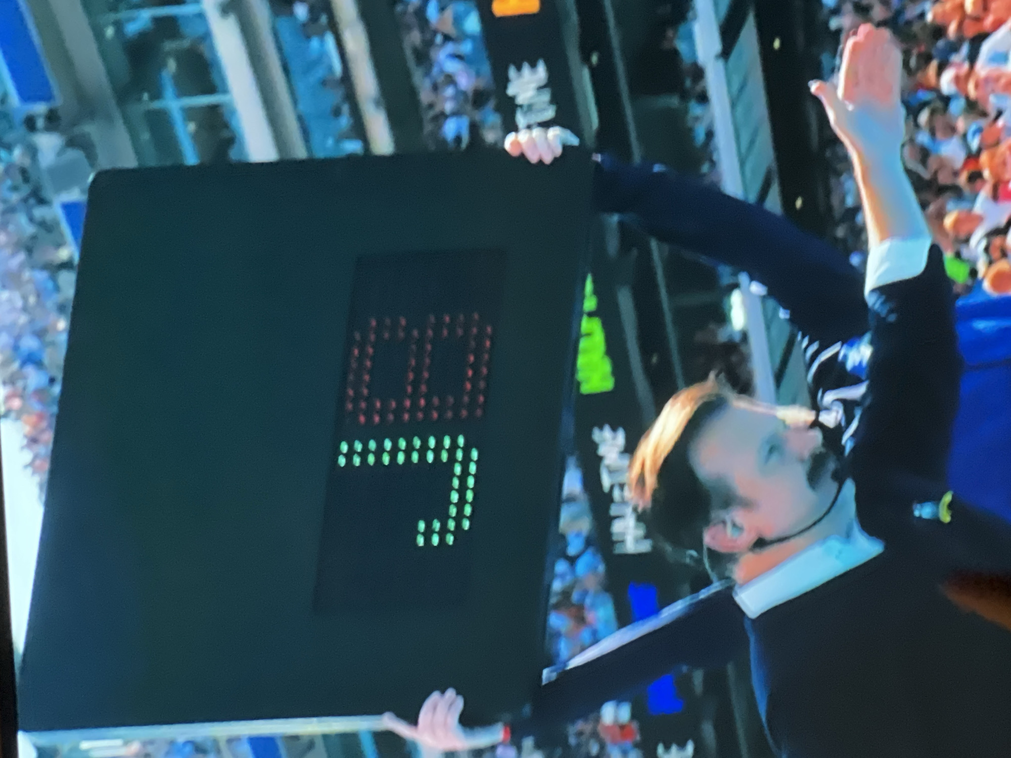 In an outdoor stadium setting, a person with a mustache and light-colored hair is holding up a large, rectangular, dark-colored electronic display board. The display board shows the letters "JB" formed by red and green LED lights. The letter "J" is rendered in green, and the letter "B" is rendered in red. Below the "JB" display, partial text is visible, including "HALFTIME" in white LED lights on a dark background, and other unintelligible text in green and white. The person holding the board is wearing a dark suit jacket over a light-colored collared shirt, and a black earpiece. Their left hand is holding the top left corner of the board, and their right arm is extended forward, with their palm open. The background shows blurred spectators in a stadium seating area.
