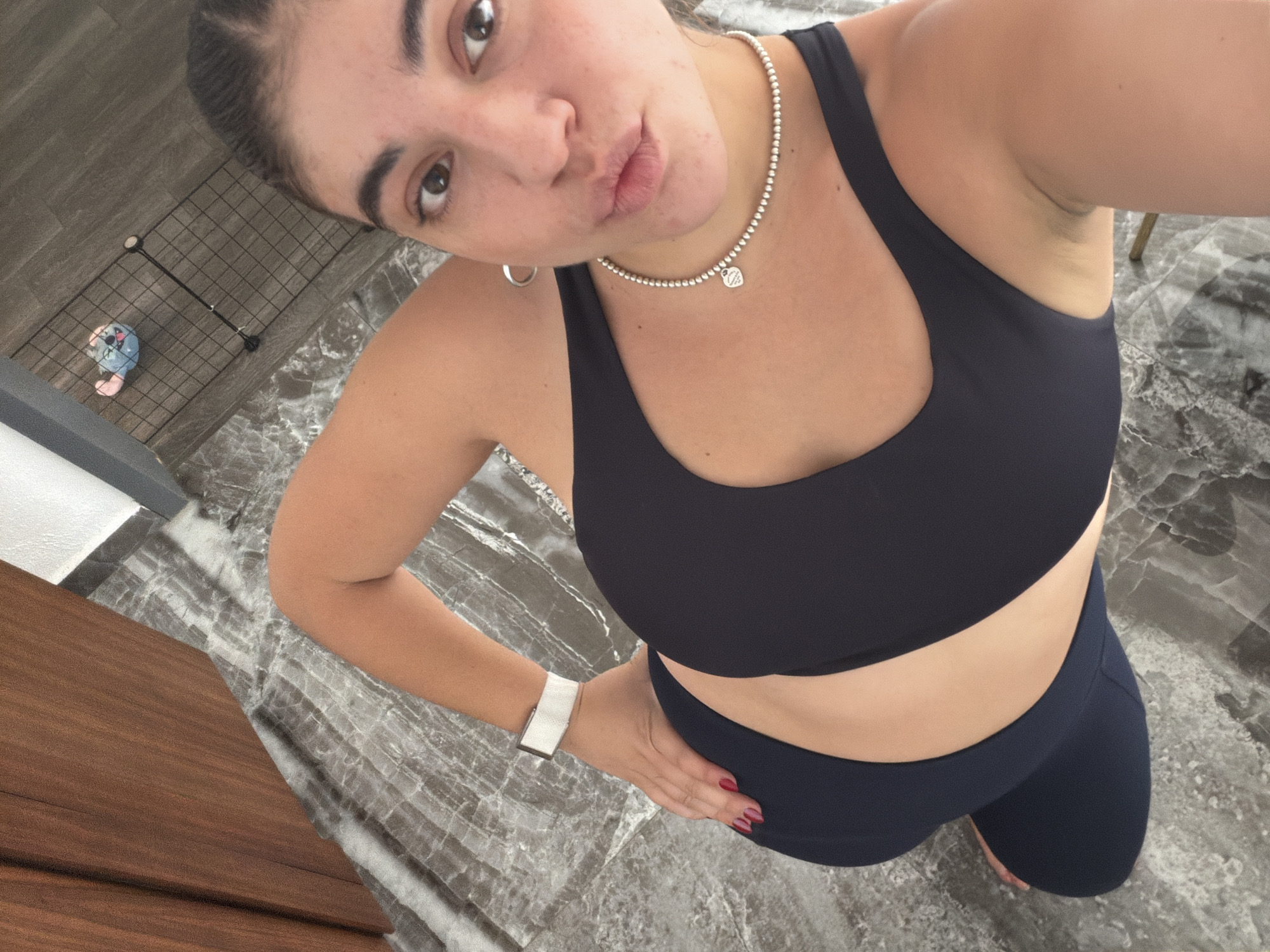 A young woman is captured in a high-angle selfie, looking directly at the camera with a slight pout. Her dark hair is pulled back, and she wears a dark-colored sports bra and matching leggings. Around her neck is a silver beaded necklace featuring a small, heart-shaped charm clearly engraved with "Tiffany & Co." She also has a silver hoop earring in her visible ear and a white smartwatch or fitness tracker on her left wrist. Her fingernails are painted red, and her left hand rests on her hip.
The setting is an indoor space, likely a room in Puebla, Mexico. The floor features a prominent grey and white marbled pattern. To the left, a section of a dark wooden surface is visible. In the background, a small, light blue and pink plush toy or doll is seen inside a black wire grid enclosure, which appears to be a baby playpen or fence. The lighting is bright and even, typical of an indoor environment, and the woman's expression suggests a confident and playful demeanor.