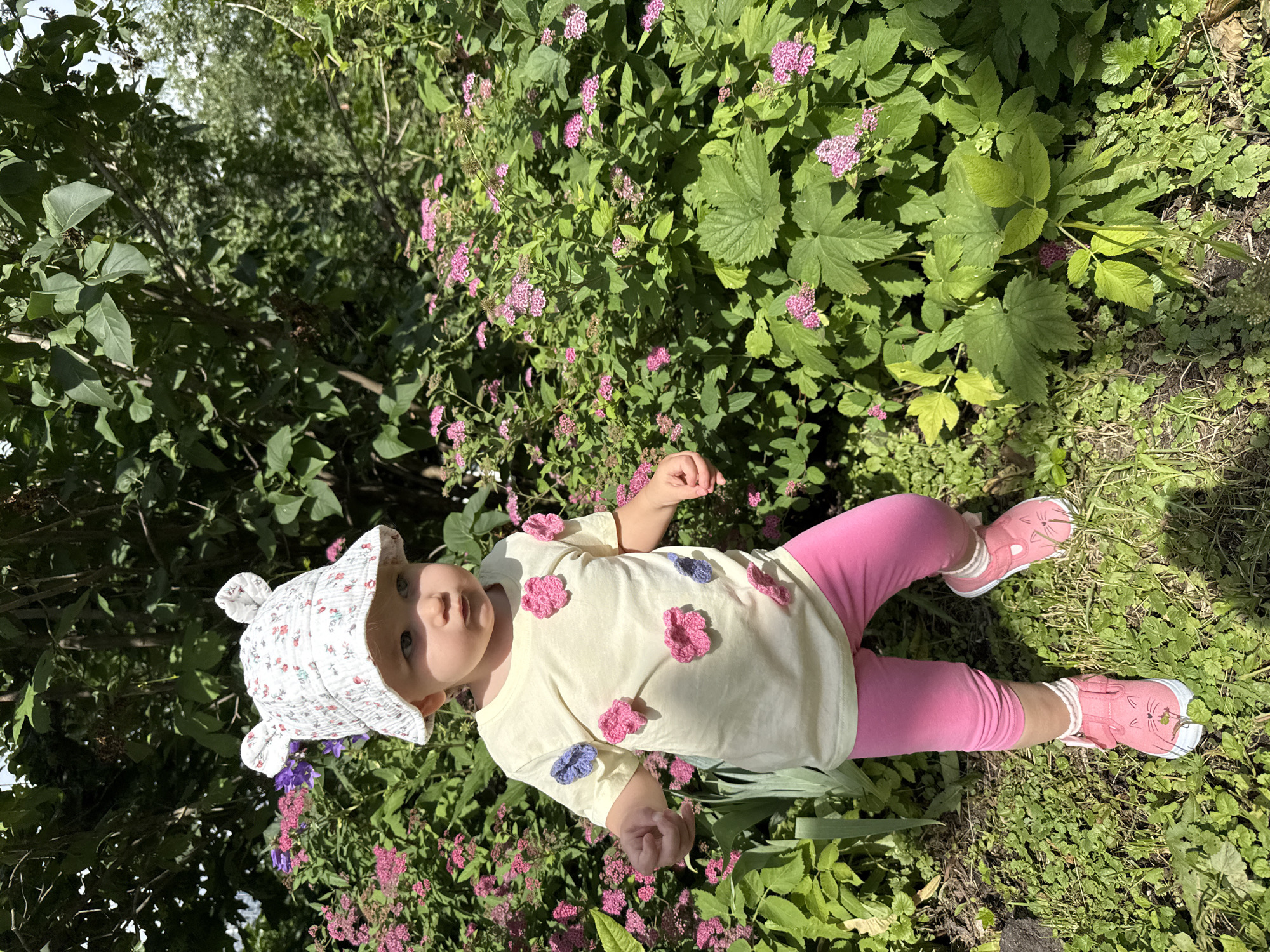 A full-body, eye-level shot shows a young toddler, approximately 1-2 years old, standing in a grassy, garden area with dense green foliage and scattered pink flowers. The toddler is wearing a white bucket hat with a floral pattern and bear ears, a light yellow t-shirt adorned with pink and blue crocheted flowers, bright pink leggings, and pink sneakers with cat face details. The toddler is looking slightly to the right with a neutral expression and appears to be walking or standing. The background consists of various green leaves and branches from trees and bushes, suggesting an outdoor setting. There are no visible indications of the location being in Moscow, Russia.