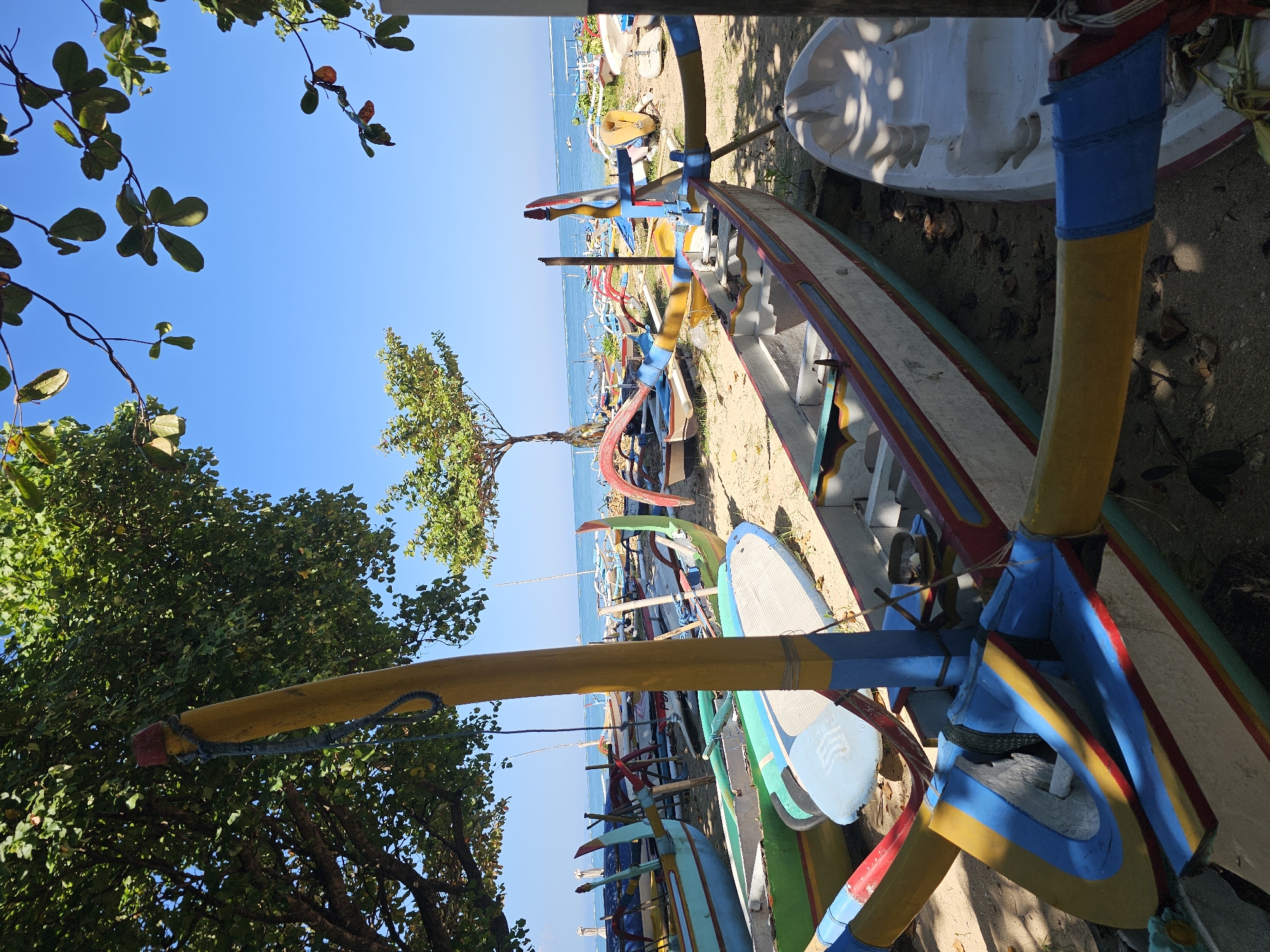 The image captures a sunny scene on a beach in Denpasar, Indonesia. The foreground is dominated by several colorful traditional Balinese fishing boats, known as jukung, resting on the sand. These boats are characterized by their outriggers and vibrant paintwork in shades of yellow, blue, red, and green. One prominent boat in the center has a yellow outrigger pole with blue and red accents. To the right, a white kayak or small boat with seat indentations is partially visible. In the mid-ground, numerous other jukung are lined up along the shore, some with their poles extended. Beyond the boats, a calm, blue ocean stretches to the horizon under a clear sky. A few distant boats and what appear to be buoys or markers are visible on the water. Trees with green foliage frame the scene from above, casting shadows on the sand and boats, suggesting it is midday. The beach itself is sandy, with some dry leaves scattered on the ground. The overall impression is one of a typical coastal scene in Bali, likely a fishing village or a popular spot for boat rentals. There are no people visible in the image.