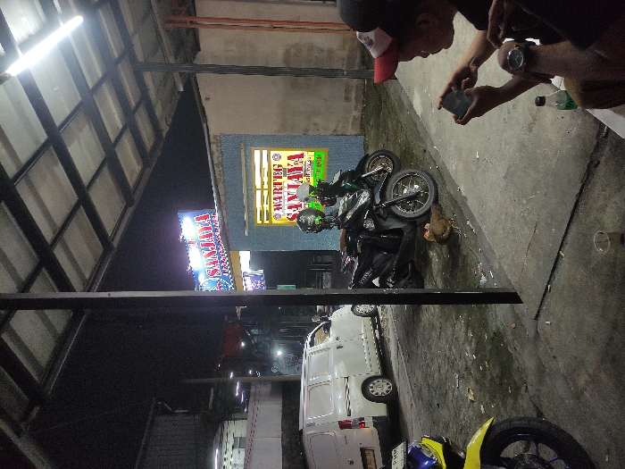 This nighttime image captures a street-side scene at Warteg Sanjaya in Bogor, Indonesia. A large blue and white "SANJAYA" sign illuminates the background, situated above a light-blue building with a prominent yellow "WARTEG SANJAYA" sign on its facade. The immediate foreground is covered by a corrugated metal roof supported by black metal pillars, lit by overhead fluorescent tubes.
To the right, a person wearing a red and white baseball cap and a dark shirt is seated, holding a smartphone horizontally with both hands, focused on its screen. A watch is visible on their left wrist. A second individual's arm is partially visible to the left of the first person. On the concrete ground near the seated figures, a clear plastic cup and a green plastic bottle are visible, along with scattered cigarette butts.
In the middle ground, two black motor scooters (one identifiable as a Honda Beat) are parked, facing left, each with a helmet hanging from its handlebars. A brown chicken is on the concrete ground near these parked vehicles. To the left, a white panel van is parked facing right. The front wheel and partial yellow and blue fender of another motorbike are visible in the extreme left foreground. The concrete ground appears worn, with visible dirt and some moist patches. A darker, possibly industrial building is present in the distant left background.