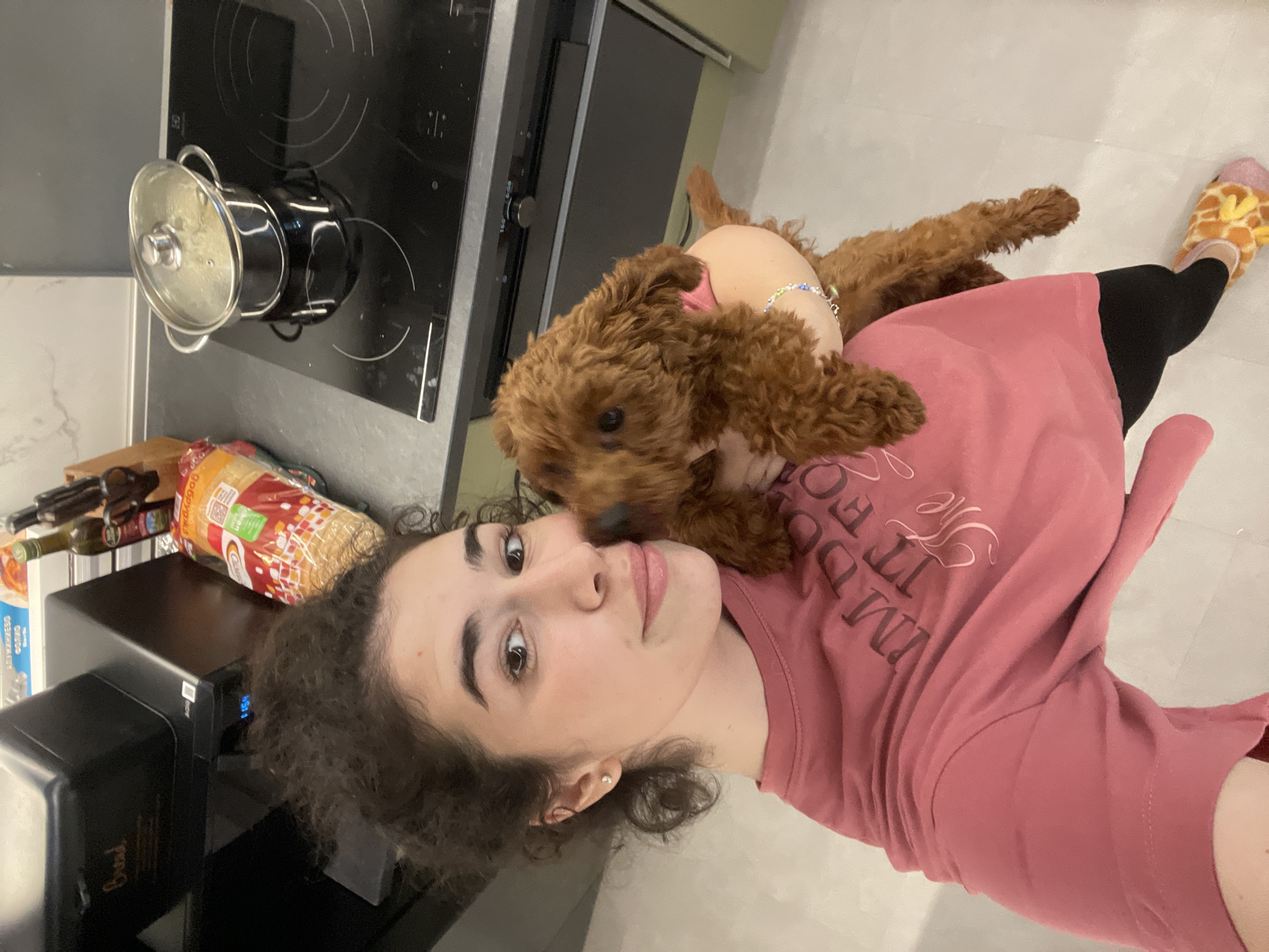 A young woman with curly dark hair and light skin is taking a selfie with a brown poodle on her shoulder. She is wearing a pink t-shirt with white lettering and black leggings. She is also wearing giraffe-patterned slippers. The poodle is wearing a pink collar and a colorful beaded necklace.
The background shows a kitchen. There is a black stovetop with a pot on it, and a black bread machine. There are also various food items and bottles on the counter. The flooring is light-colored tile.
There is no information to determine the location is the city of Sofia, Bulgaria.