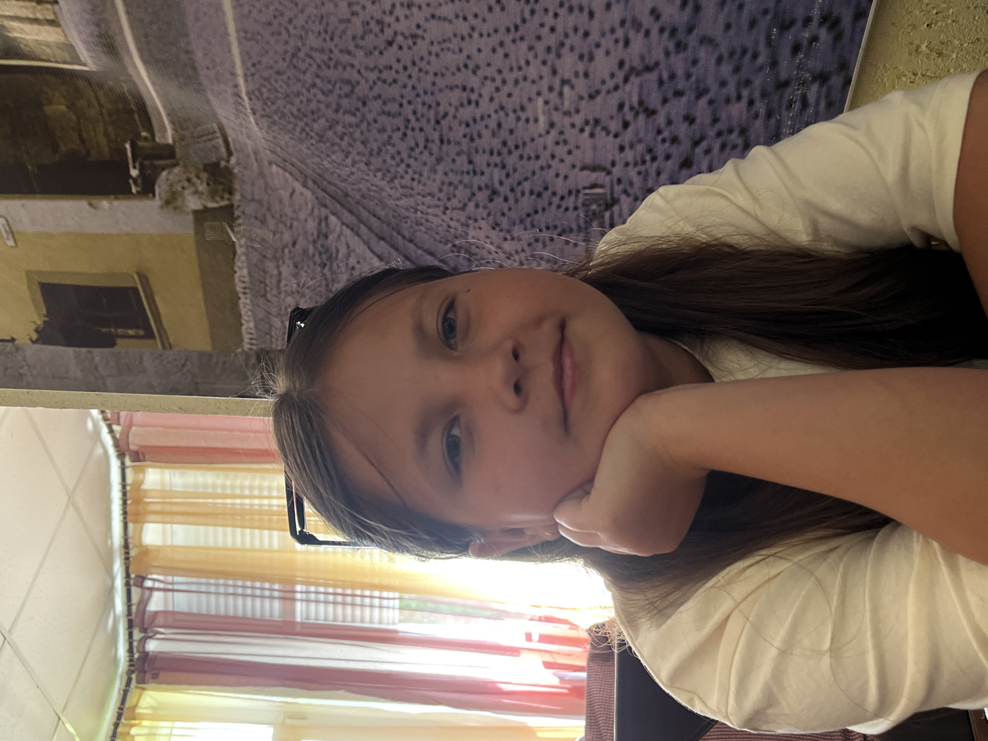 A young female subject, appearing to be in her pre-teen years, is depicted indoors in a medium close-up, located in Novopokrovskaya, Russia. She has long, straight, dark brown hair and wears a light cream or off-white short-sleeved t-shirt. Dark-framed eyeglasses are perched on top of her head. She faces the camera directly with a neutral expression, resting her chin on her right hand, which is supported by an unseen surface.
The background is visually divided. To the subject's left, a window is covered by multi-colored sheer curtains in shades of yellow, orange, and reddish-pink, allowing diffused natural light to enter. A white grid-patterned acoustic tile ceiling is visible in the upper left. To the subject's right, a wall is adorned with a large mural or tapestry depicting an antiquated street scene. Features of the mural include a yellow building with a dark, recessed window and a textured, cobblestone-like ground rendered in shades of grey and purple. The scene suggests a casual indoor setting.