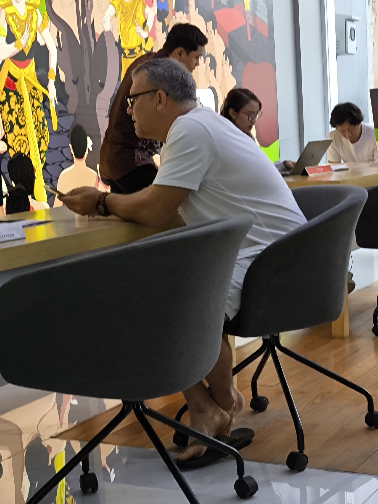 The photograph depicts a group of people in what appears to be a modern workspace, possibly in North Kuta, Indonesia. A man with short grey hair wearing glasses and a white t-shirt is seated at a light-colored table, looking to his left and holding a smartphone. He is wearing black flip-flops. Another person wearing a brown patterned shirt stands behind him. Further down the table, a woman with glasses is using a laptop, and a man in a white shirt sits next to her.
The setting is a room with a vibrant, artistic mural on the wall behind the group. The table has some papers and objects on it. The foreground features a grey office chair, with parts of other chairs visible. The floor is shiny and reflects the light, and there is a window in the background.