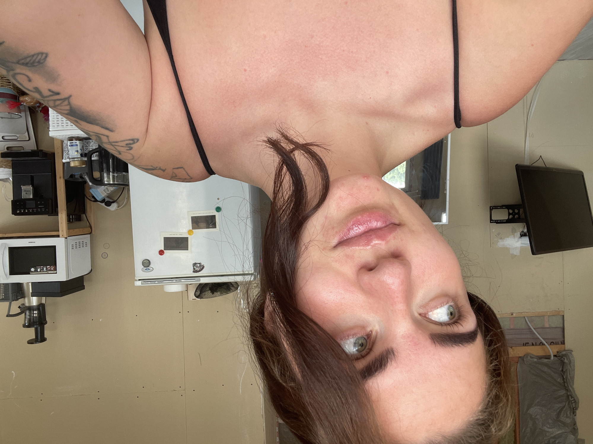 A young woman with brown hair and green eyes is posing for a selfie in what appears to be a construction or unfinished room. She is wearing a black spaghetti strap top. The background includes a white refrigerator with magnets on it, a microwave on a shelf, a coffee machine, a black computer monitor mounted on the wall, and some construction materials like insulation and drywall. The room has unpainted walls and exposed framing in some areas. The woman is looking off to the side with a slight smile.