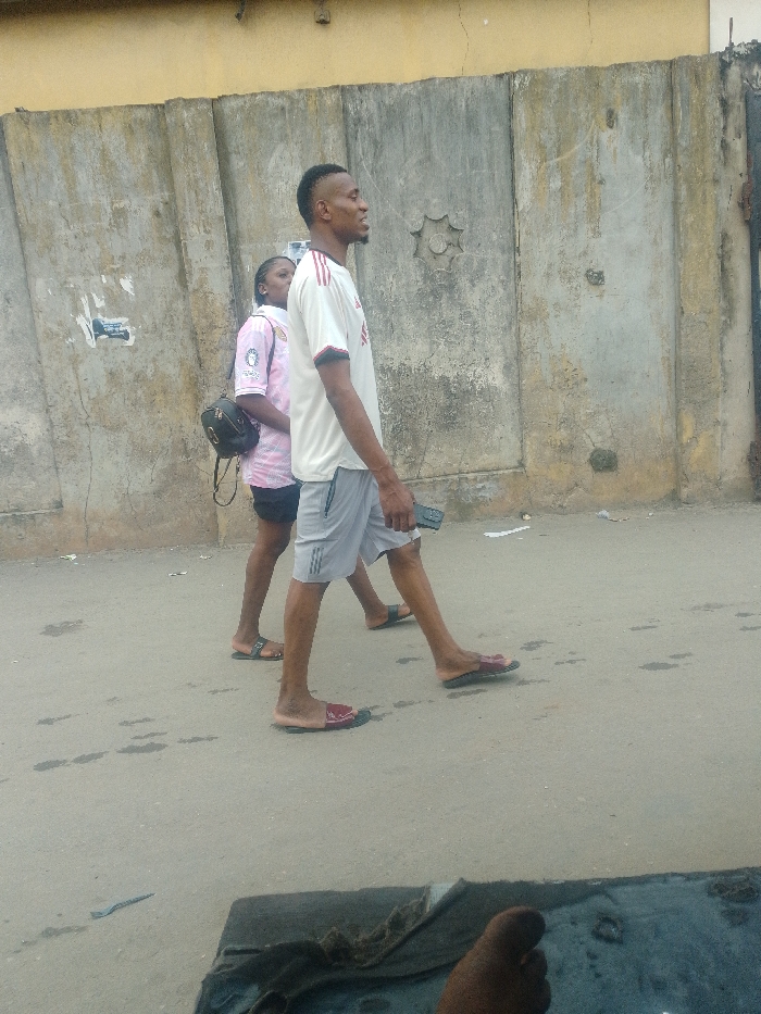 The image depicts an outdoor street scene in Agani, Nigeria, featuring two individuals walking from left to right past a textured wall.
In the foreground, a male individual with dark skin and short, faded hair walks in profile. He wears a white short-sleeved athletic shirt with dark stripes on the shoulders, light grey shorts with darker stripes, and dark-red slides. He holds a dark object, possibly a mobile phone, in his right hand. His gaze is directed slightly upwards and forward.
Behind and slightly to his left, a female individual with dark skin walks in profile. She wears a pink and white patterned short-sleeved shirt, dark shorts, and dark brown slides. A small black backpack is slung over her left shoulder.
The background consists of a weathered grey concrete wall, showing discoloration, stains, and remnants of torn paper. A star-shaped decorative element is embedded in the wall. Above this concrete section, a strip of a lighter, beige-colored wall is visible. The ground is a grey paved surface with various dark stains. In the bottom right corner, a bare human foot is partially visible, suggesting the presence of a third, unidentifiable individual, possibly seated.
