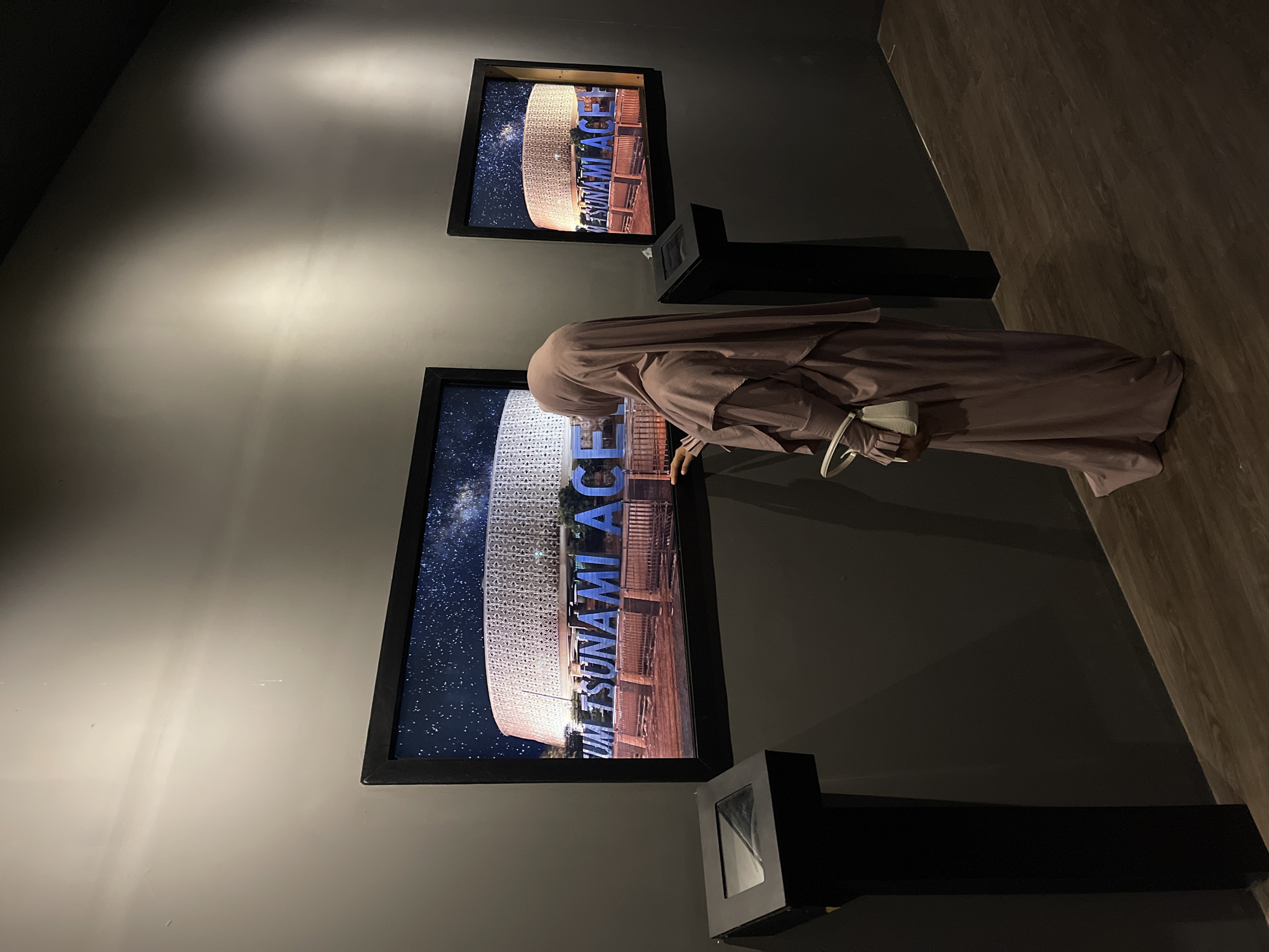 A woman wearing a light purple hijab and dress stands in front of a large framed photograph. The photograph displays a night sky filled with stars and a large, modern building with intricate geometric patterns. In front of the building, large blue letters spell out "UM TSUNAMI ACEH". The woman is facing away from the camera and appears to be touching the photograph. To the left of the photograph is a black pedestal with a glass-covered display. To the right of the woman, another smaller framed photograph of the same scene is visible. The walls are dark gray, and the floor is wooden. The scene appears to be within a museum or exhibition space.