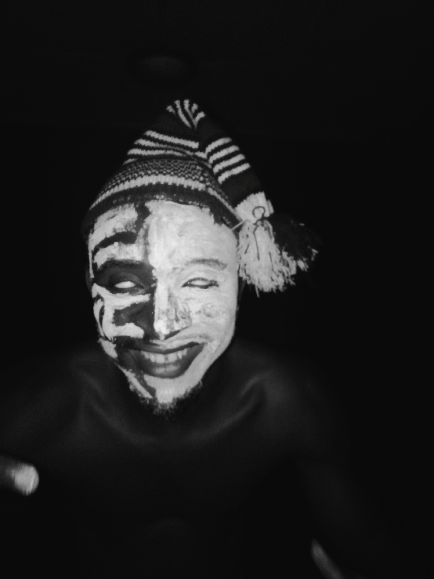 The image is a black and white close-up of a person with partially painted white face and dark eye sockets, grinning. The person is wearing a striped wool cap. There is no information about the location being in Onitsha, Nigeria or any information of city in the provided image.