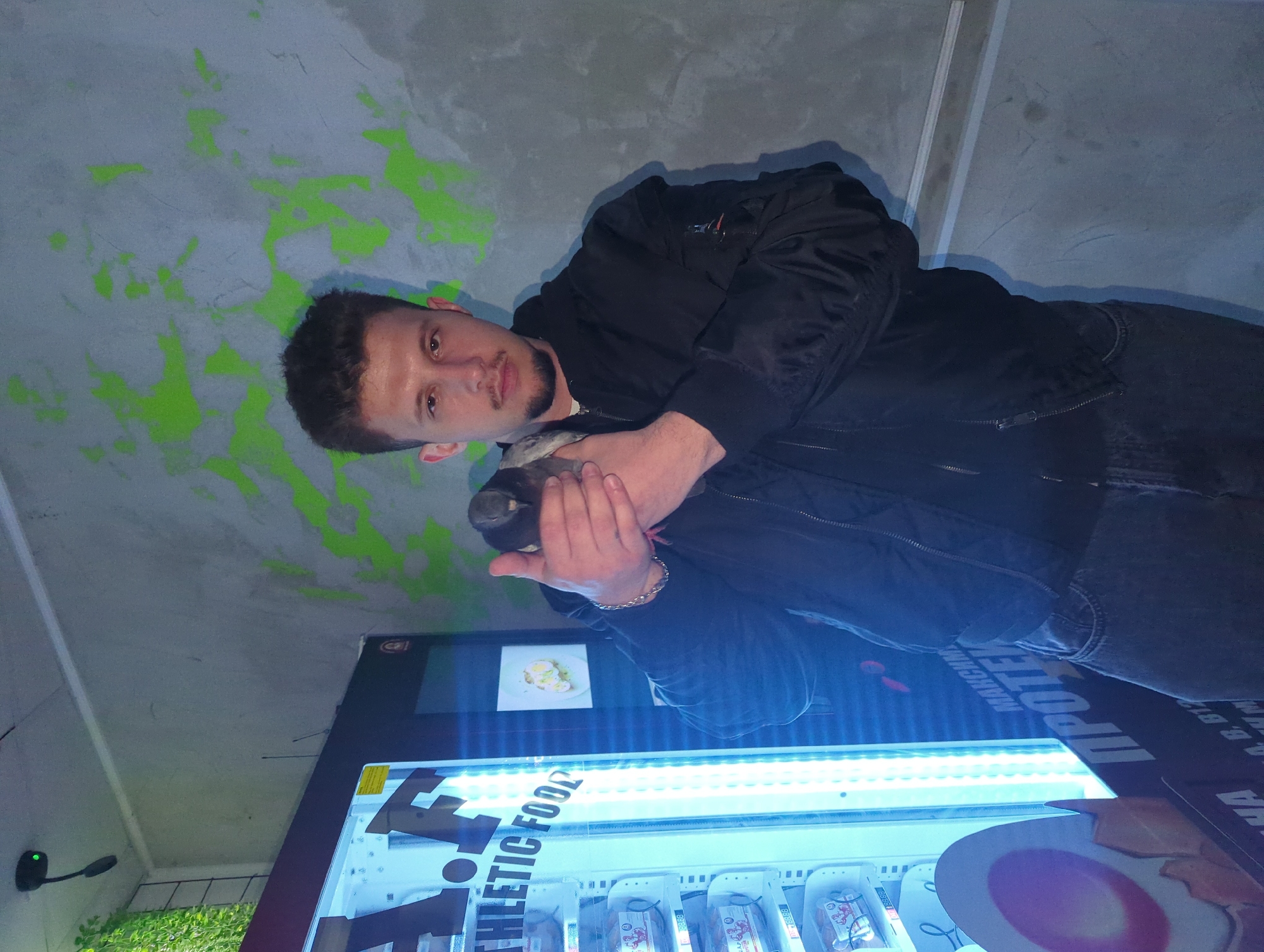 The image, taken in Razgrad, Bulgaria, shows a young man standing in an indoor setting in front of a brightly lit vending machine. The man, who appears to be in his twenties, has short dark hair, a light mustache, and a short beard. He is wearing a black bomber jacket over a lighter top and dark trousers. He holds a dark-colored bottle or cylindrical object with both hands in front of his chest, looking directly at the camera with a neutral expression. A silver-colored chain bracelet is visible on his right wrist.
The prominent vending machine is illuminated by strong blue-white lights, highlighting the products inside. A digital display at the top shows an image of food, possibly eggs with greens. The main section displays packaged items, likely healthy snacks or protein products, with some Cyrillic text on their labels. The words "ATHLETIC FOOD" are clearly visible in white letters on the vending machine's front panel. Partially visible Cyrillic text includes "ПРОТЕК" and "МАКСИМАЛНА" on the machine's side panels.
The background features a light grey, textured wall, possibly concrete, adorned with abstract splatters of neon green paint. A black lighting fixture with a small green LED indicator is mounted on the wall to the lower left. The overall lighting suggests an indoor space, possibly a gym or a modern facility, where the vending machine serves as a focal point. The man appears to be posing for a photograph.