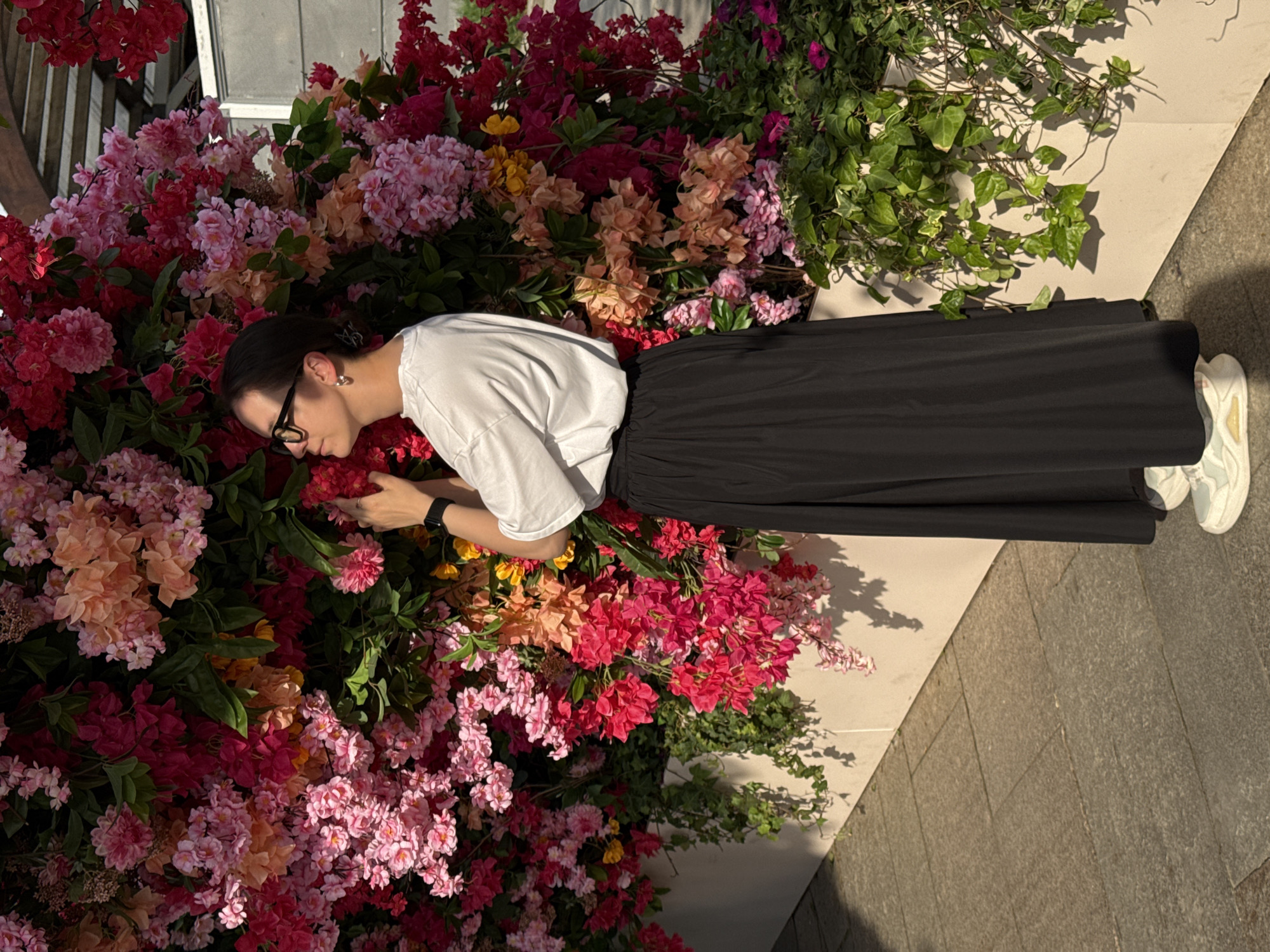 A person lies among an arrangement of various colorful flowers and green plants. The individual, wearing glasses, a white shirt, and a long black skirt, is positioned on a light-colored surface next to a grey brick walkway. The lighting suggests the scene is outdoors, possibly in a garden area within Moscow, Russia, given the plants and flower arrangement, and it is a daytime scene. The person seems to be interacting with the flowers.