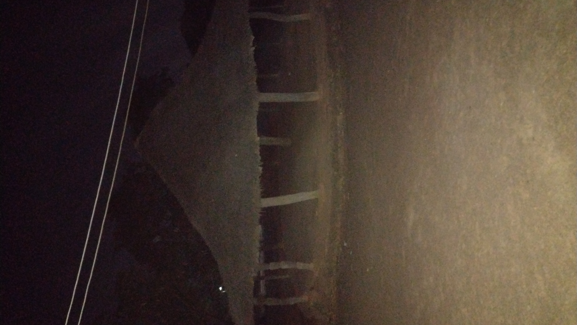 The image depicts a dimly lit, unpaved area at night. A section of what appears to be a concrete or paved surface runs vertically on the right side of the image. Along the left of that surface, there is an uneven edge with what appear to be remnants of structures, possibly broken or unfinished steps or ledges. To the left of this area, the scene is predominantly dark with two parallel lines of light, possibly wires, running diagonally from upper left to lower center. The overall scene is poorly lit, with limited details discernible, and the location in Jos, Nigeria, is not immediately evident from the image itself. There are no people visible in the frame.