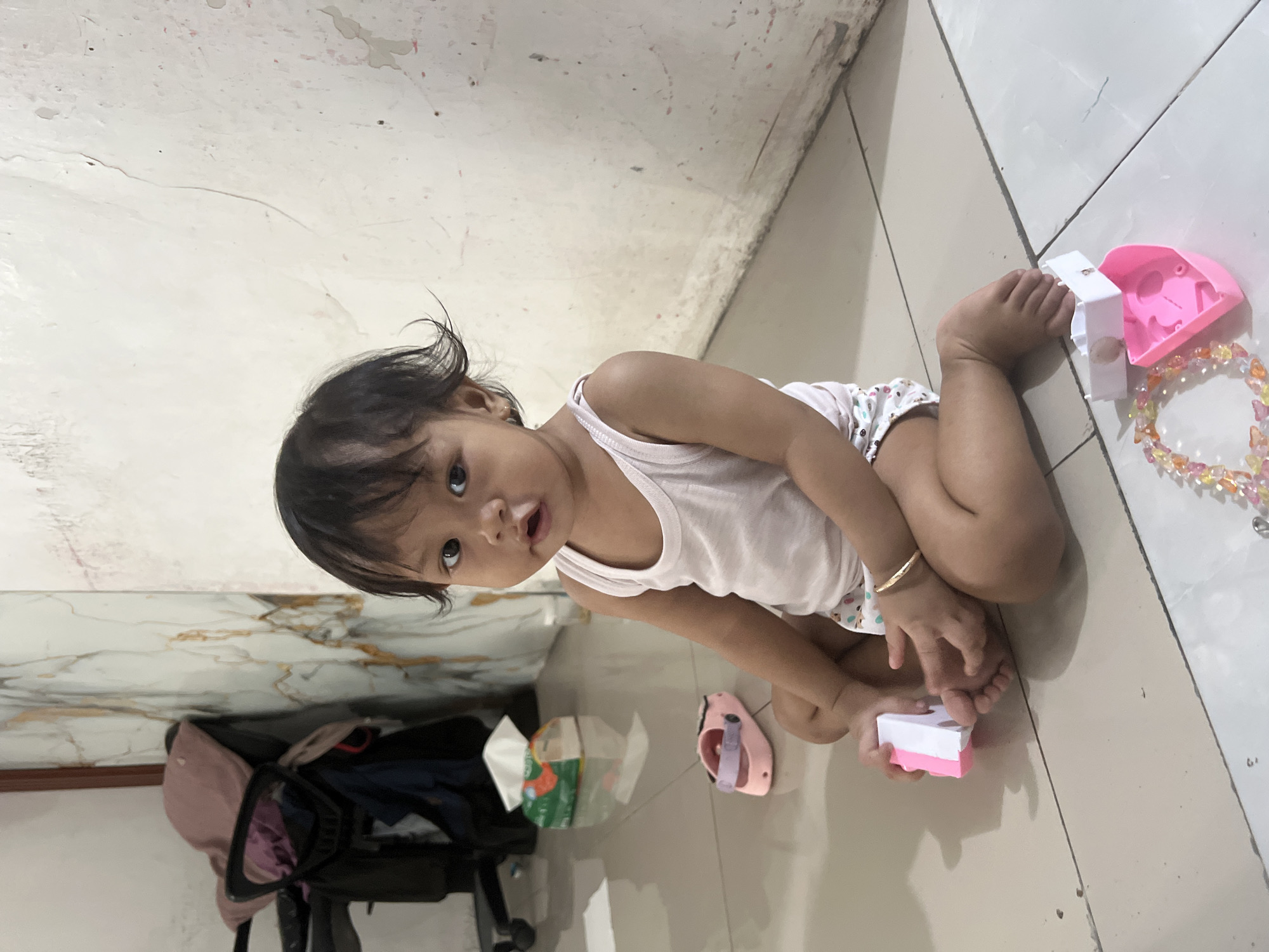 A young child sits on a tiled floor, playing with toys. The child is wearing a white tank top and patterned shorts. In front of the child are two pink toy houses, a bracelet made of colorful beads, and a pink shoe. A box of tissues and a stroller are visible in the background. The room appears to be in a home located in Kecamatan Pati, Indonesia. The walls are a neutral color with some discoloration and scuff marks.