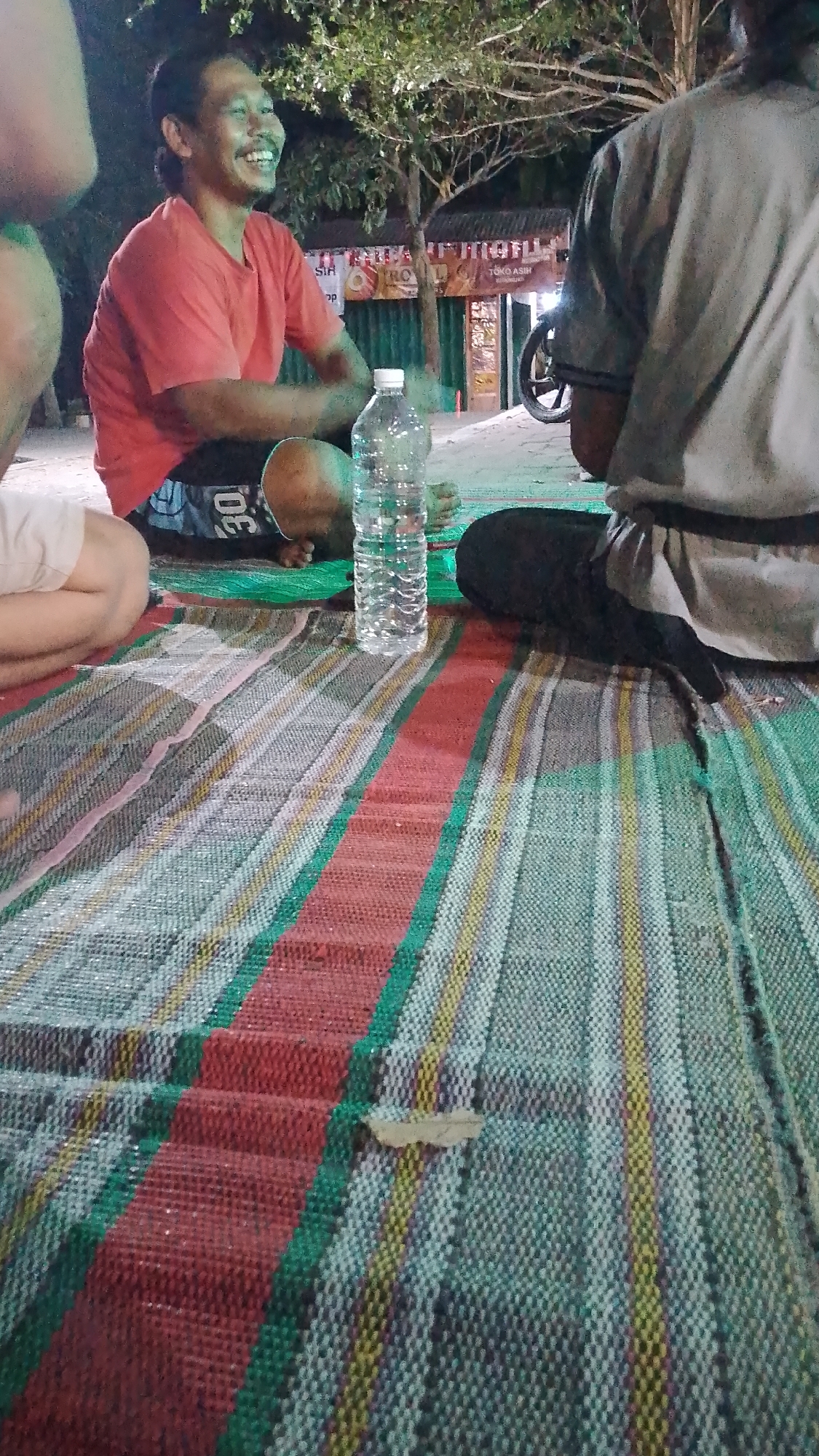 The image shows a group of people sitting on a mat on the ground, likely outdoors at night. A man in a red t-shirt is smiling and looking to his left, with his hands resting in his lap. He is wearing black shorts with the number "30" visible. Next to him is a plastic bottle of water. To his right, a person wearing a grey t-shirt and dark pants is visible from the back. The ground is covered with a colorful mat with stripes of red, green, yellow, and white. In the background, trees and a building with signage are visible, suggesting a commercial or public area in Karanglangu, Indonesia. The lighting suggests it is evening or nighttime.