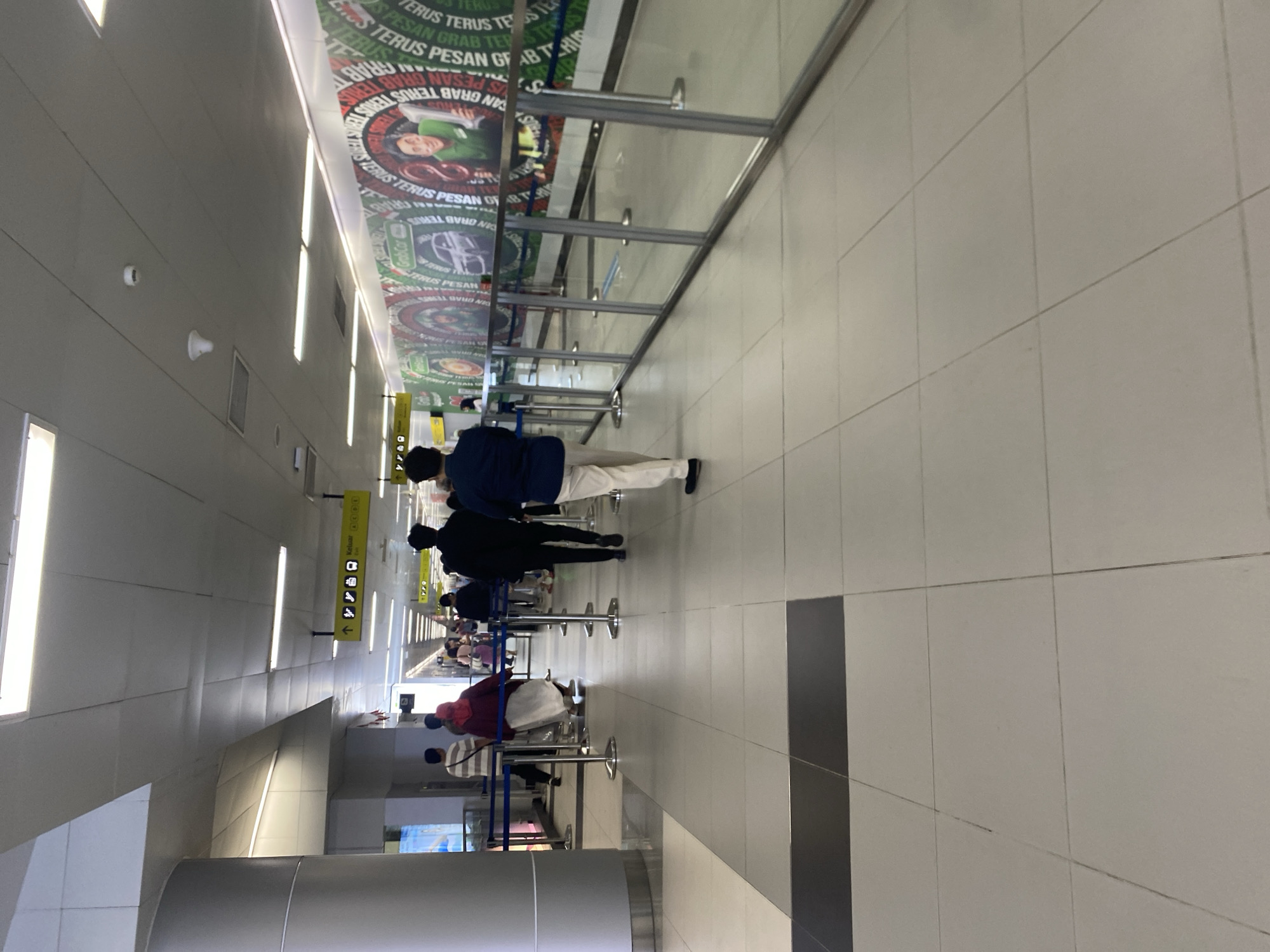 The image depicts an interior scene within a public building, likely an airport terminal, judging by the presence of directional signs pointing to "Keluar" (Exit) and symbols for transportation. The environment is clean and modern, with tiled floors and a suspended ceiling equipped with fluorescent lighting. A long, transparent glass barrier with metal stanchions divides the walkway, indicating a queuing or waiting area.
Several individuals are present in the scene, most of them walking away from the viewer or standing in line. On the left side of the walkway, a group of people is visible. One person is wearing a striped t-shirt and dark pants, another a maroon headscarf and white skirt, and a third is partially obscured. In the center of the frame, two men are walking away. The man in the foreground is wearing a dark jacket and light-colored pants, while the man behind him is wearing a black jacket. Further down the walkway, more people are seen in the distance, also appearing to be in a queue.
The walls of the building are adorned with large, colorful posters or banners, one of which features circular designs and text that appears to be in Indonesian, with the word "GRAB" prominently displayed. This suggests a commercial advertisement, possibly for a ride-sharing service, common in urban transportation hubs. The overall impression is one of orderly movement within a busy public space. Given the signage and the likely context of such an environment, this scene is consistent with a location in Central Jakarta, Indonesia.