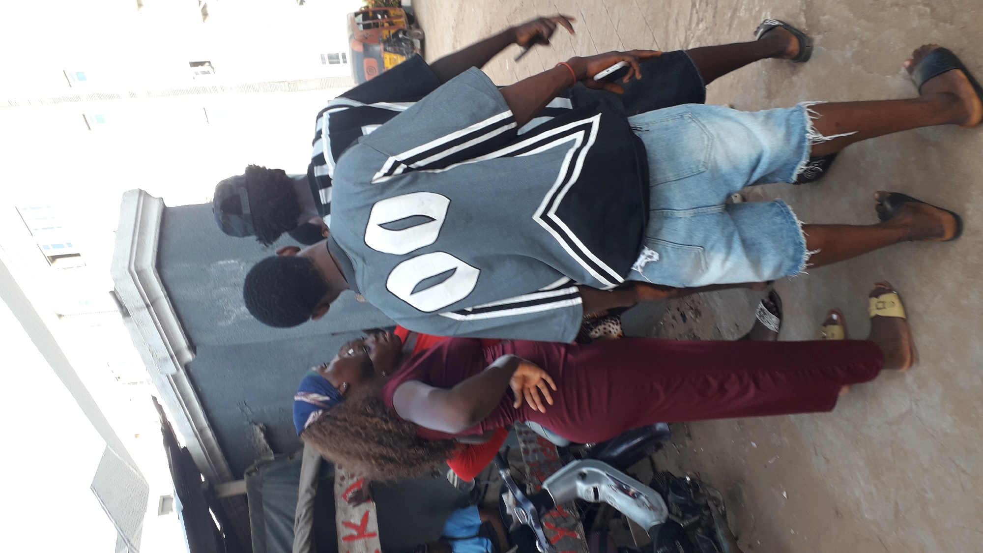 The image shows three people standing outside in Nkpor, Nigeria. Two men are standing close together; the man closest to the camera is wearing a gray jersey with the number 00 on the back, denim shorts, and black sandals. The second man is wearing a black shirt and blue denim shorts. A woman is standing close to the two men, wearing a maroon dress. The ground is concrete. A tricycle can be seen in the background.