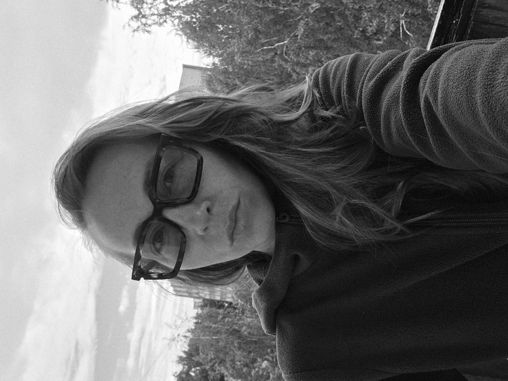 A monochrome close-up portrait depicts an adult female looking directly at the camera with a neutral expression. She has long, wavy hair, visible on both sides of her face and draped over her shoulders. Her face displays visible freckles across the nose and cheeks. She wears large, rectangular, dark-framed glasses with apparently tinted lenses. Her attire consists of a dark-toned (appearing black or very dark grey in monochrome) fleece jacket with a visible front zipper and an unzipped collar. The texture of the fleece fabric is distinct.
The background indicates an outdoor setting, possibly an elevated position such as a balcony or terrace in an urban or suburban environment, in the city of Unknown, Unknown. Dense deciduous trees and foliage are visible immediately behind her and to her right, with the bare branches of a tree prominent on the far right. Beyond the trees, several indistinct building structures are visible under an overcast sky filled with clouds. A dark, textured surface, possibly a railing or ledge, is visible at the bottom right corner of the frame.
