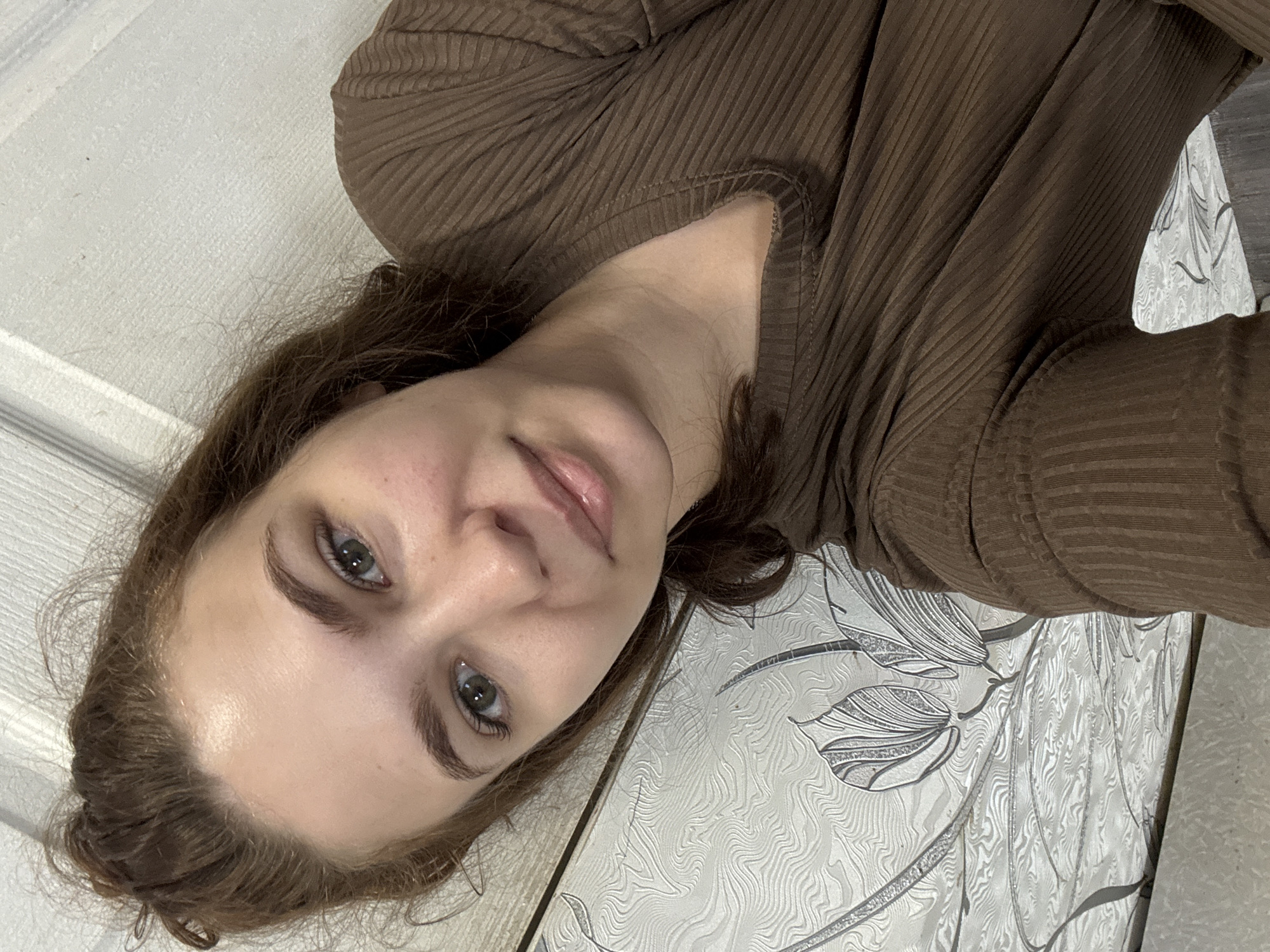 A young woman with fair skin, light-brown hair, and green eyes is pictured in a close-up, tilted headshot. She is wearing a brown ribbed long-sleeve shirt. The background consists of a white surface with a patterned design, possibly a floral or textured wallpaper, and a white door in the distance. There is no indication of the location being Nizhnyaya Gar', Russia. The photo does not provide forensic details or information about actions or interactions.
