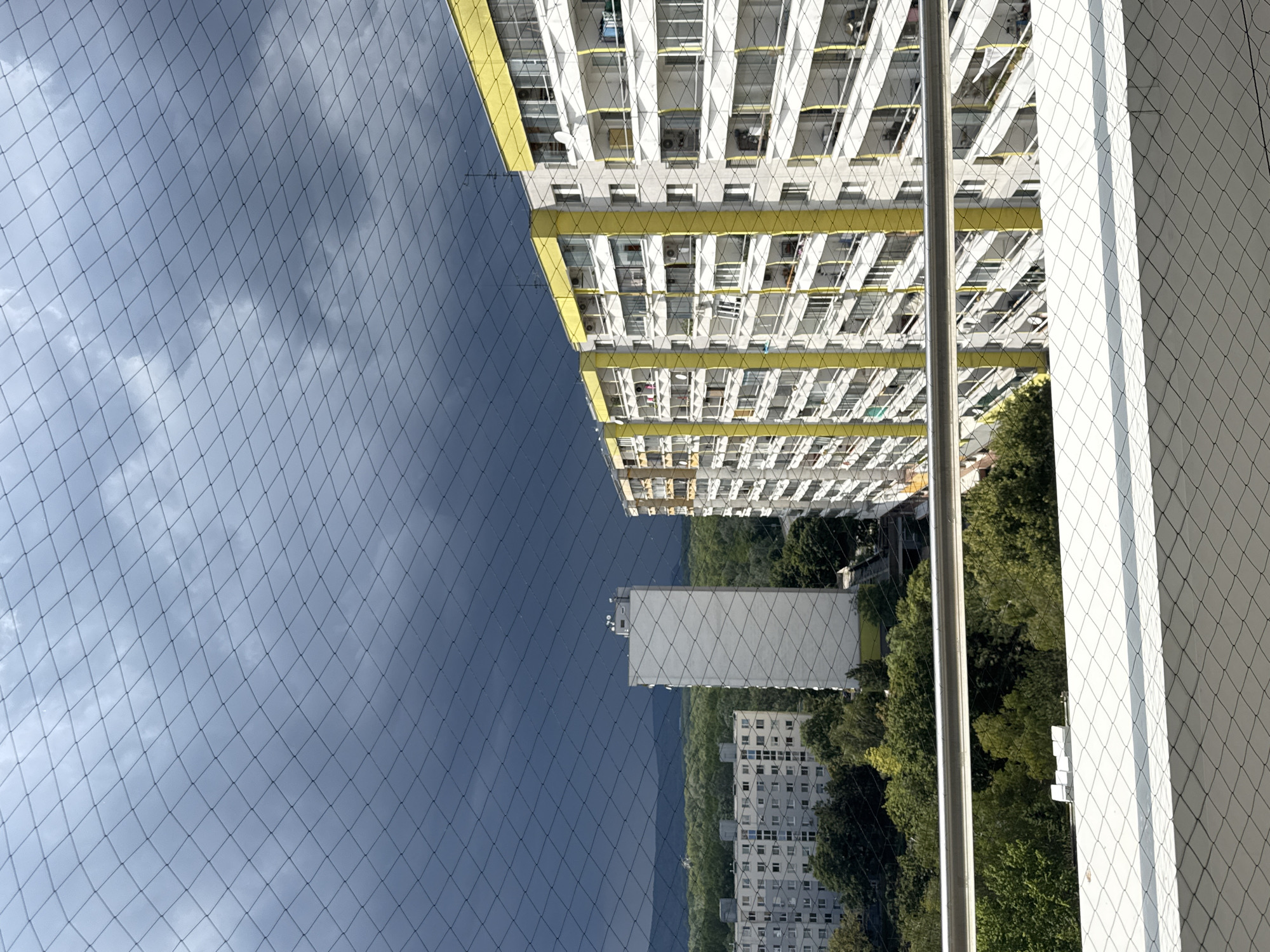 The image captures a view from a balcony in Bratislava, Slovakia, looking out at a cityscape under a stormy, dark sky. A fine, black mesh, likely a bird deterrent net, spans the foreground and mid-ground, creating a diamond pattern that partially obscures the view. In the mid-ground, several apartment buildings are visible. To the right, a tall, yellow and white building with numerous balconies dominates the scene. To the left, a long, white apartment building with many windows stretches across the frame. Between these two structures, a slender, white tower stands prominently. The background features a dense forest and rolling hills, partially visible beneath the ominous clouds. The overall impression is one of urban development contrasted with the approaching storm.