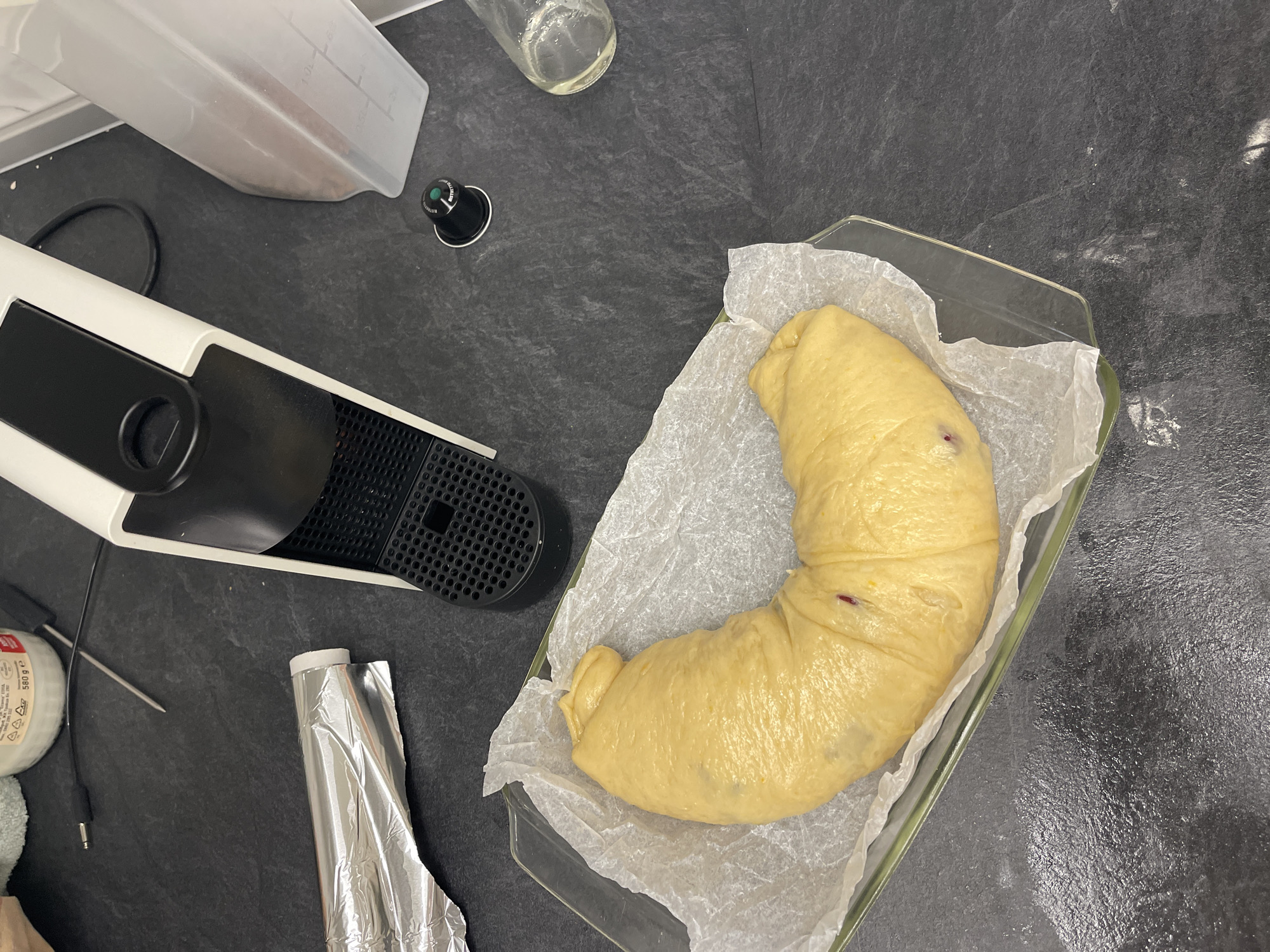 The image shows a kitchen counter scene. On the counter, there's a dough prepared for baking in a glass container lined with baking paper. Nearby are a white Nespresso coffee machine, a clear plastic container, a glass bottle, a used Nespresso capsule, foil, and a temperature probe next to a container labeled "580 g e." The setting appears to be a modern kitchen with a dark countertop. The image does not contain information that would indicate a location in Sofia, Bulgaria.