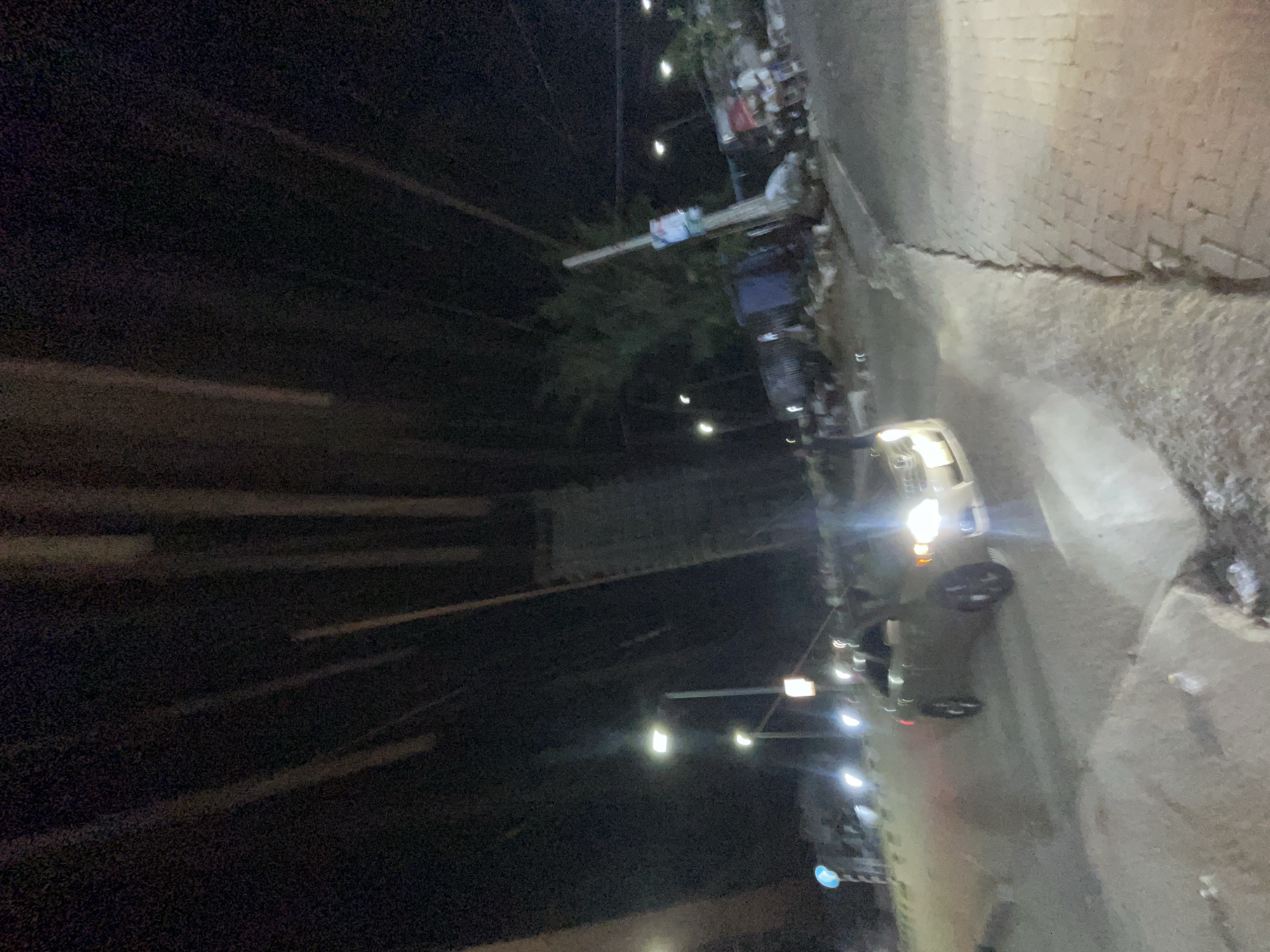 At night in Ilado, Nigeria, a street scene unfolds. The road is wet, reflecting the glare of vehicle headlights. A vehicle with illuminated headlights is parked near the roadside. Streetlights cast a dim glow on the surroundings. One or more pedestrians appear to be in close proximity to the parked vehicle. A sidewalk runs along the edge of the road, lined with buildings. Overhead power lines are visible.