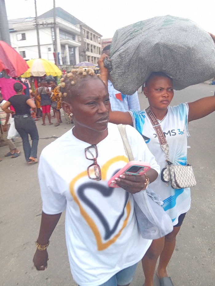 The scene depicts an outdoor street or market environment in Ura Nta Umuarandu, Nigeria, under diffused daylight conditions. In the immediate foreground, two women are walking forward.
The woman on the left, closer to the camera, possesses a dark complexion and short, blonde, bunched hair. She wears a white short-sleeved t-shirt featuring a large graphic of a heart outlined in yellow and black. Brown-framed eyeglasses rest on her shirt's neckline. Her left wrist displays multiple gold-colored bracelets. She holds a pink-cased smartphone in her right hand and carries a light grey shoulder bag over her left shoulder. Her expression is neutral.
Behind and slightly to the right of the first woman, a second woman with a dark complexion and short, dark hair carries a large, grey, possibly mesh bag balanced on her head, with her hands assisting its stability. She wears a light blue and white short-sleeved t-shirt with text including "LUODEN ENGDAISIA" and "MS 3½". A patterned light-colored cross-body bag with an attached circular pouch is visible across her torso. Her gaze is directed forward-right.
The background reveals a bustling urban setting. To the left, several market stalls are present, indicated by red and yellow umbrellas or awnings, beneath which various individuals are visible. One figure in a black t-shirt and black trousers is seen walking away from the camera. In the distant background, multiple multi-story buildings line the street; one prominent structure features a green roof and a lighter facade. Utility poles and overhead wires are also discernible. The ground appears to be a paved street or pathway.