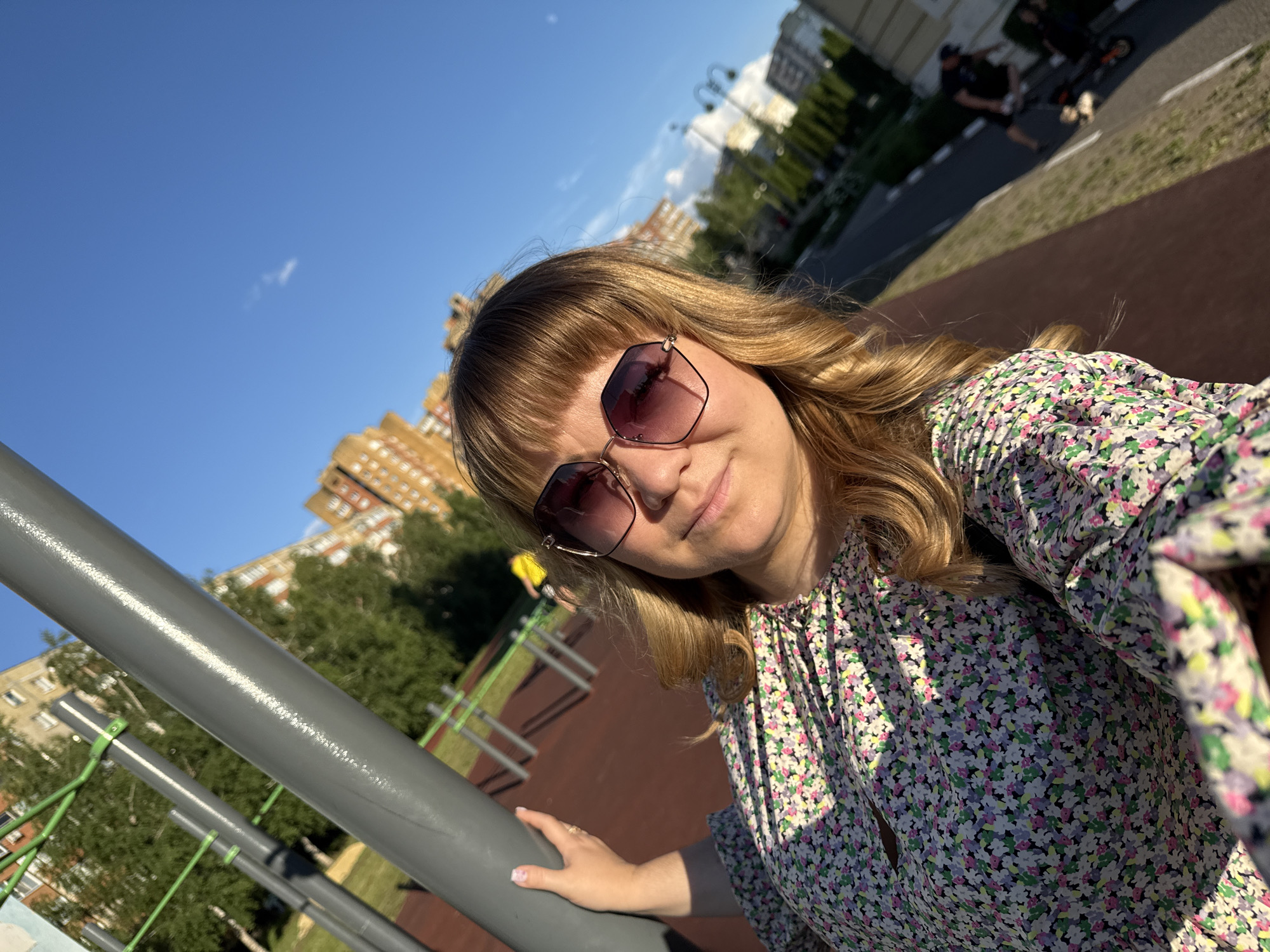 This is a selfie taken by a woman with blonde hair and bangs, wearing oversized sunglasses and a floral print dress. She is outdoors, with a clear blue sky above and buildings and trees in the background. The setting appears to be a public park or recreational area, with playground equipment visible, including a metal bar in the foreground that the woman is holding onto. The lighting suggests it is daytime, possibly late afternoon due to the warm sunlight casting shadows. The woman's expression is relaxed and happy. There are other people and scooters in the background, indicating a public space with activity. The location is Stary Oskol, Russia.