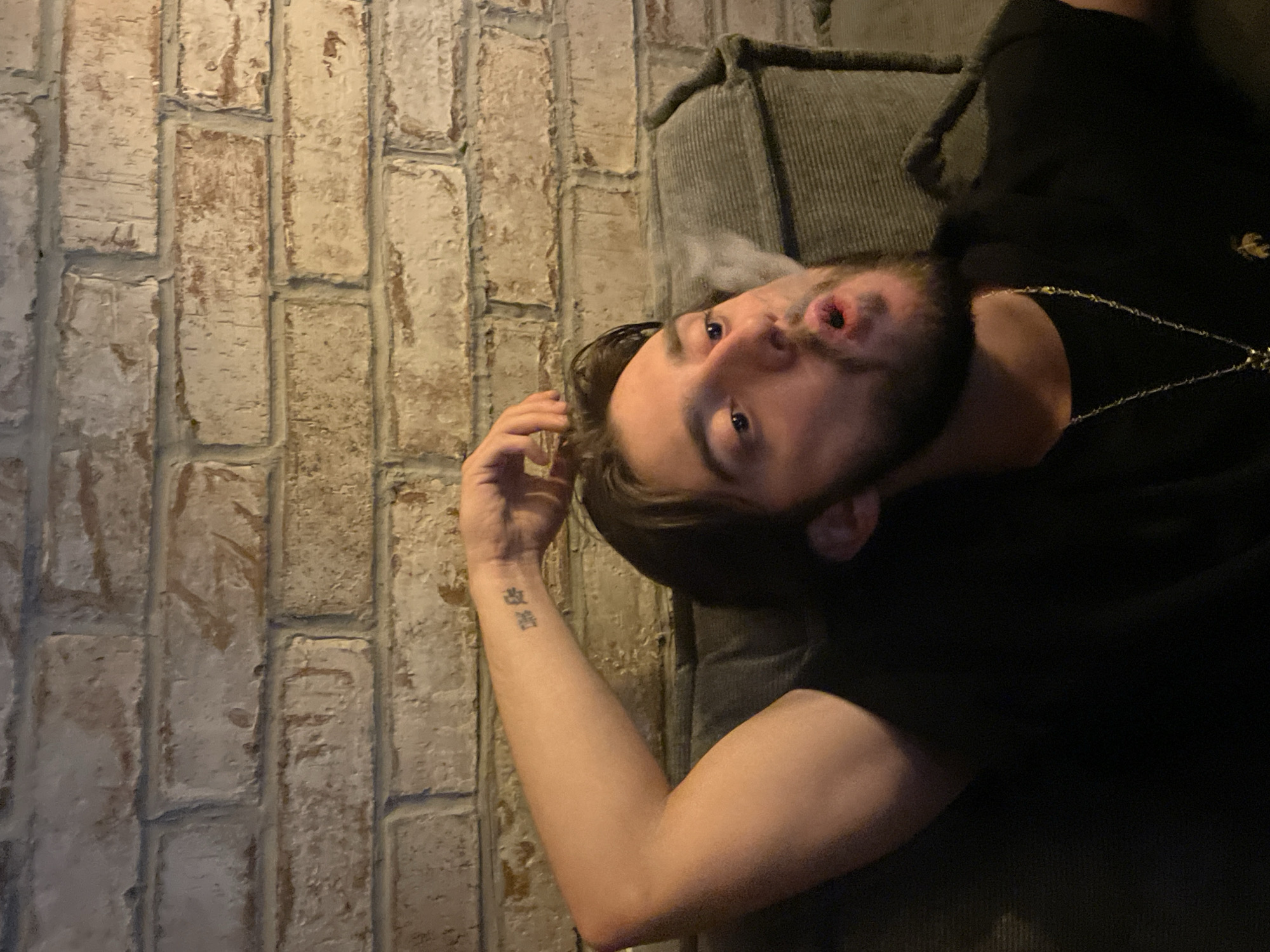 The image shows a man with a beard and dark hair exhaling smoke, possibly from a cigarette or vape, in front of a brick wall. He is wearing a black t-shirt and a necklace. The man is leaning back on what appears to be a couch or armchair. The background is a textured brick wall. There is a tattoo on his left forearm. The lighting is dim, suggesting an indoor setting.