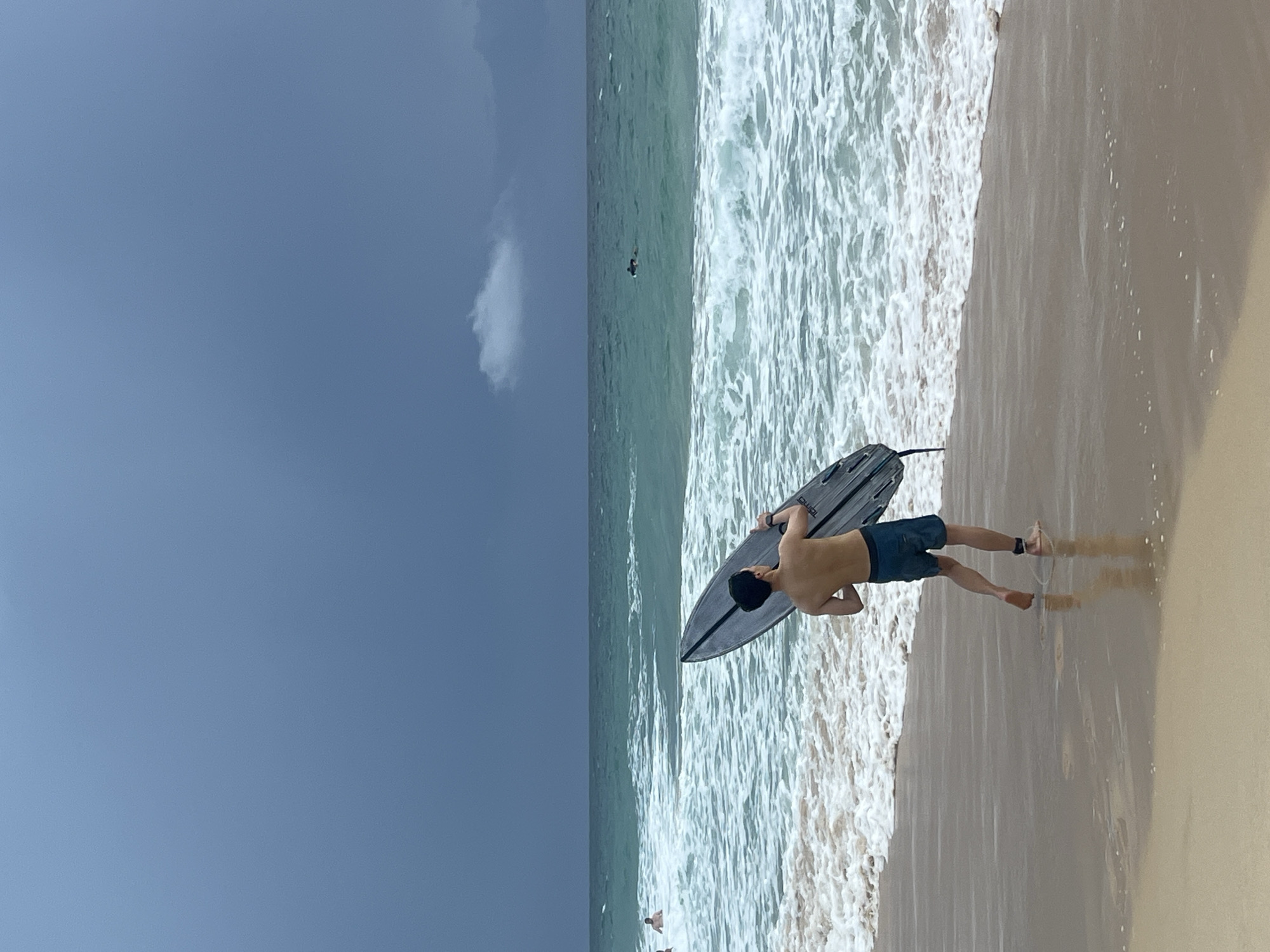 A man, viewed from behind, stands on a sandy beach holding a black bodyboard. He is wearing blue shorts. The man is facing the ocean, with waves crashing onto the shore. The ocean water is a greenish-blue color, and the sky is a deep, overcast blue with a single, small white cloud visible. The overall atmosphere appears to be cloudy, suggesting it might be an overcast day, possibly late afternoon or early morning. The scene is set on a beach, with the wet sand reflecting the sky and the man. In the distance, a few other people are visible in the water, appearing as small figures. The lighting is subdued, consistent with cloudy weather. There is no visible text in the image. The setting is likely Choeng Thale, Thailand, as indicated by the context provided.