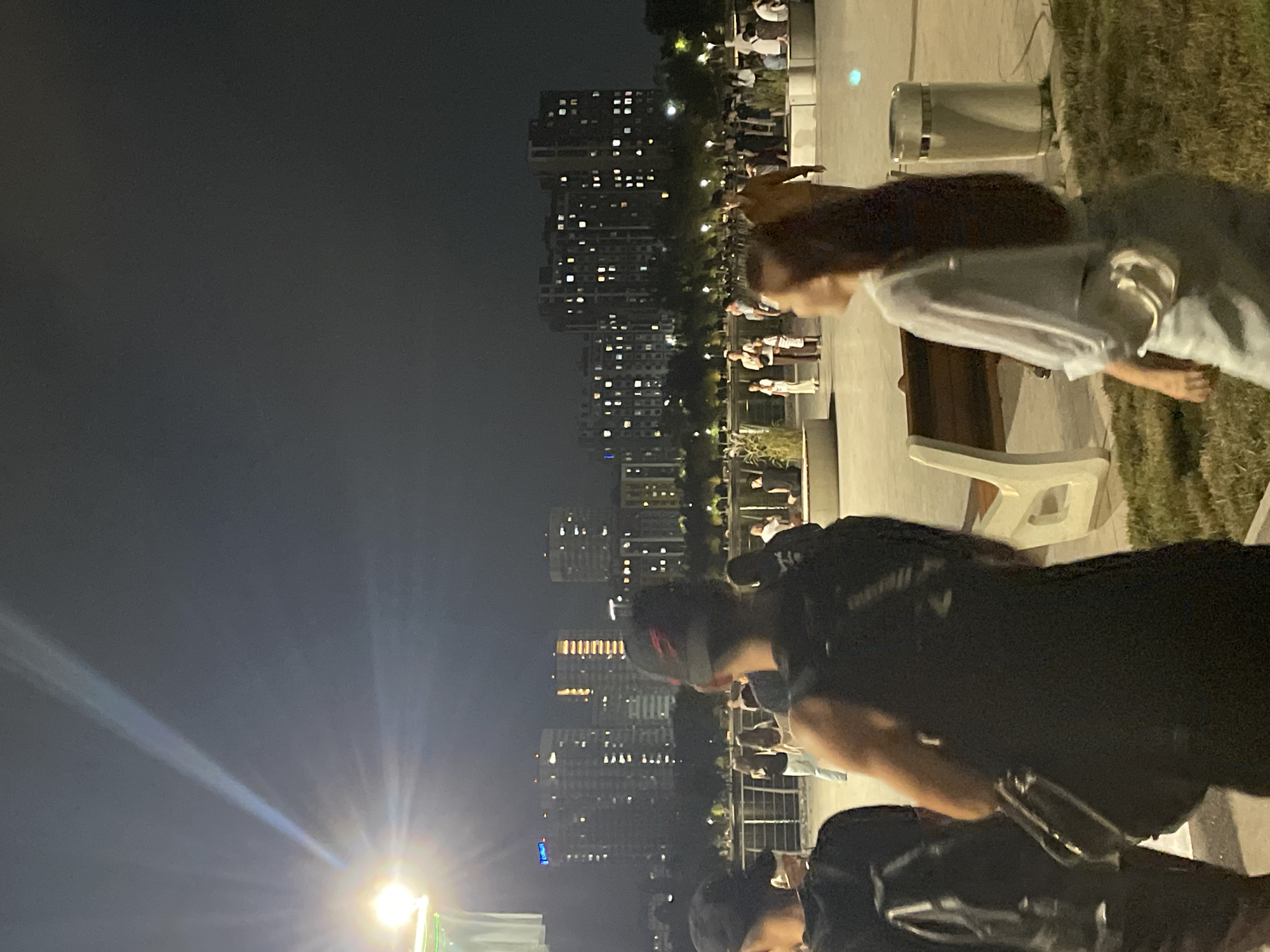 An image captured at night in Almaty, Kazakhstan, shows a group of people in an outdoor public space with a city skyline in the background. The foreground is dominated by the backs of three individuals: two men wearing dark clothing and baseball caps, and a woman with long dark hair wearing a light-colored top and light pants, with a small crossbody bag. One of the men has a backpack on his left shoulder and a strap from another bag crossing his chest. The woman in the foreground is looking to her right.
In the mid-ground, several other individuals are visible, including a man in a mustard-colored shirt walking towards the right, and groups of people standing and sitting. There are also benches and what appear to be planters with tall grass or reeds. The background features a body of water, possibly a river or lake, with trees lining its edge. Beyond the trees, numerous tall apartment buildings are illuminated with many lit windows, forming the Almaty city skyline. The lighting suggests it is nighttime, with a bright light source, possibly a spotlight or stadium light, originating from the upper left of the image and casting a beam across the dark sky. The overall impression is of a lively urban park or promenade in the evening.
