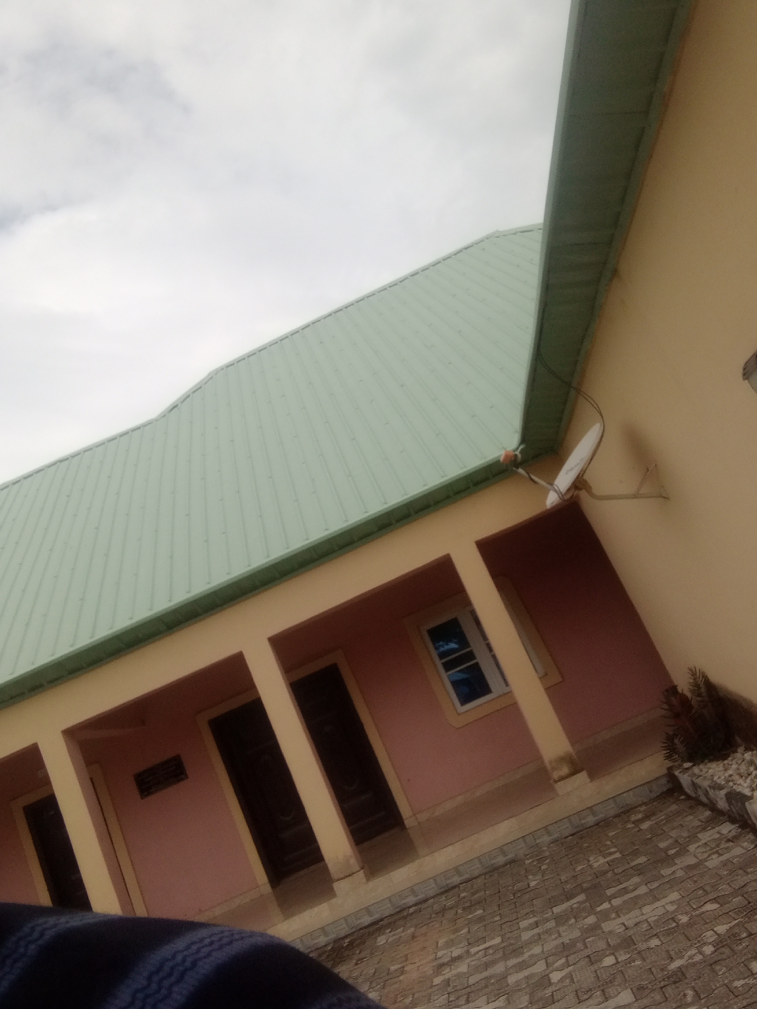 The image captures an exterior view of a building in Jalingo, Nigeria. The building features a light peach-colored wall and a light green, ribbed metal roof. A covered walkway or veranda runs along the front of the building, supported by several square columns painted in a light peach color that matches the wall. Dark brown doors are visible within the covered area, and a small window with blue tinted glass is also present. A satellite dish is mounted on the wall above the veranda. The ground in front of the building is paved with interlocking grey and tan bricks. To the right, a planter contains some dry, spiky vegetation and decorative stones. A portion of a person's shoulder and arm, wearing a dark blue and white striped garment, is visible in the lower left corner of the frame, suggesting the photo was taken from a low angle, looking up. The sky above is overcast and appears bright, indicating it is daytime. There is no discernible activity taking place, and the overall impression is of a typical building exterior on an overcast day. A small, rectangular sign with dark text is mounted on the wall between two columns.