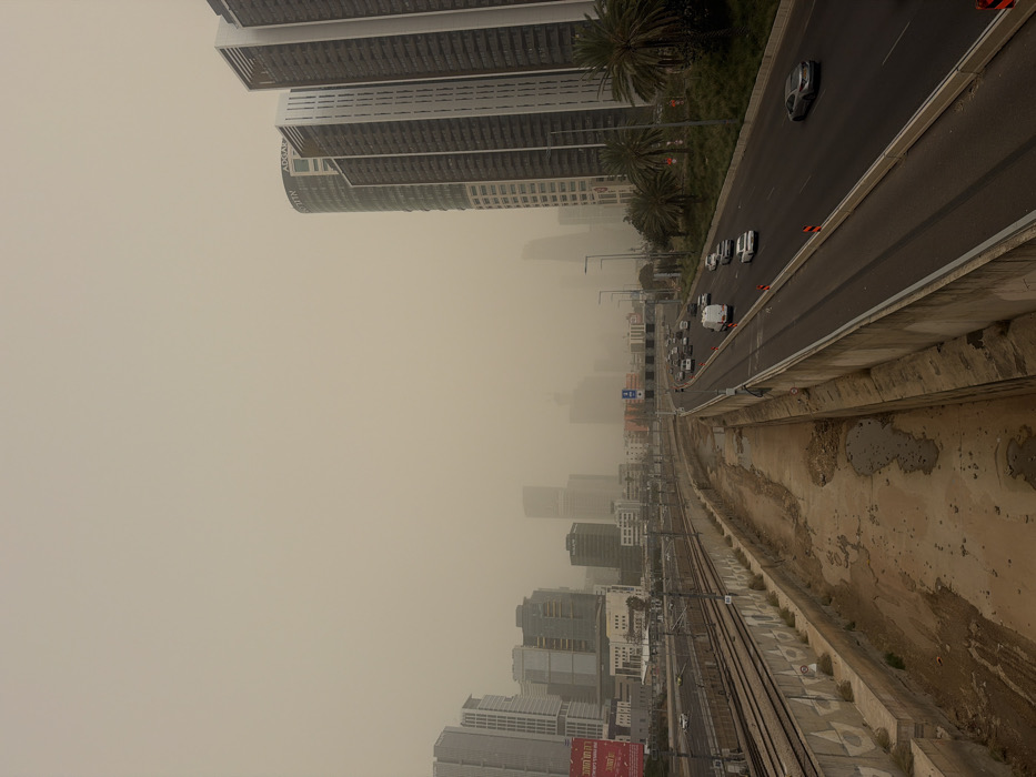The image depicts an urban landscape under a severe sandstorm or dust storm, casting an ochre-brown hue across the sky and significantly reducing visibility. The scene is located along the Ayalon Highway in Tel Aviv-Yafo, Israel, characterized by its multiple lanes, adjacent railway tracks, and surrounding high-rise buildings.
In the foreground, a concrete barrier separates the viewer's perspective from a dry, unpaved channel with muddy patches and some standing water on the left, running parallel to railway tracks. Two sets of railway tracks with overhead catenary systems are visible on a concrete embankment. Further left, a cluster of modern high-rise buildings, including one with a red billboard displaying Hebrew text, are partially obscured by the dust.
To the right, a multi-lane highway, carrying vehicles in both directions, occupies the midground. Several cars and at least one white van are present on the road; traffic appears moderate. Orange traffic cones are visible along the highway's right edge. A landscaped median strip with palm trees and green foliage separates the highway from a line of prominent, modern high-rise buildings on the far right. One of these buildings, a tall, curved structure, clearly displays "ADGAR" near its top. Other adjacent buildings feature distinctive vertical dark and light paneling. The cityscape extends into the distance, with numerous structures blurred and rendered indistinct by the heavy atmospheric dust. No individuals are visible outside of vehicles.