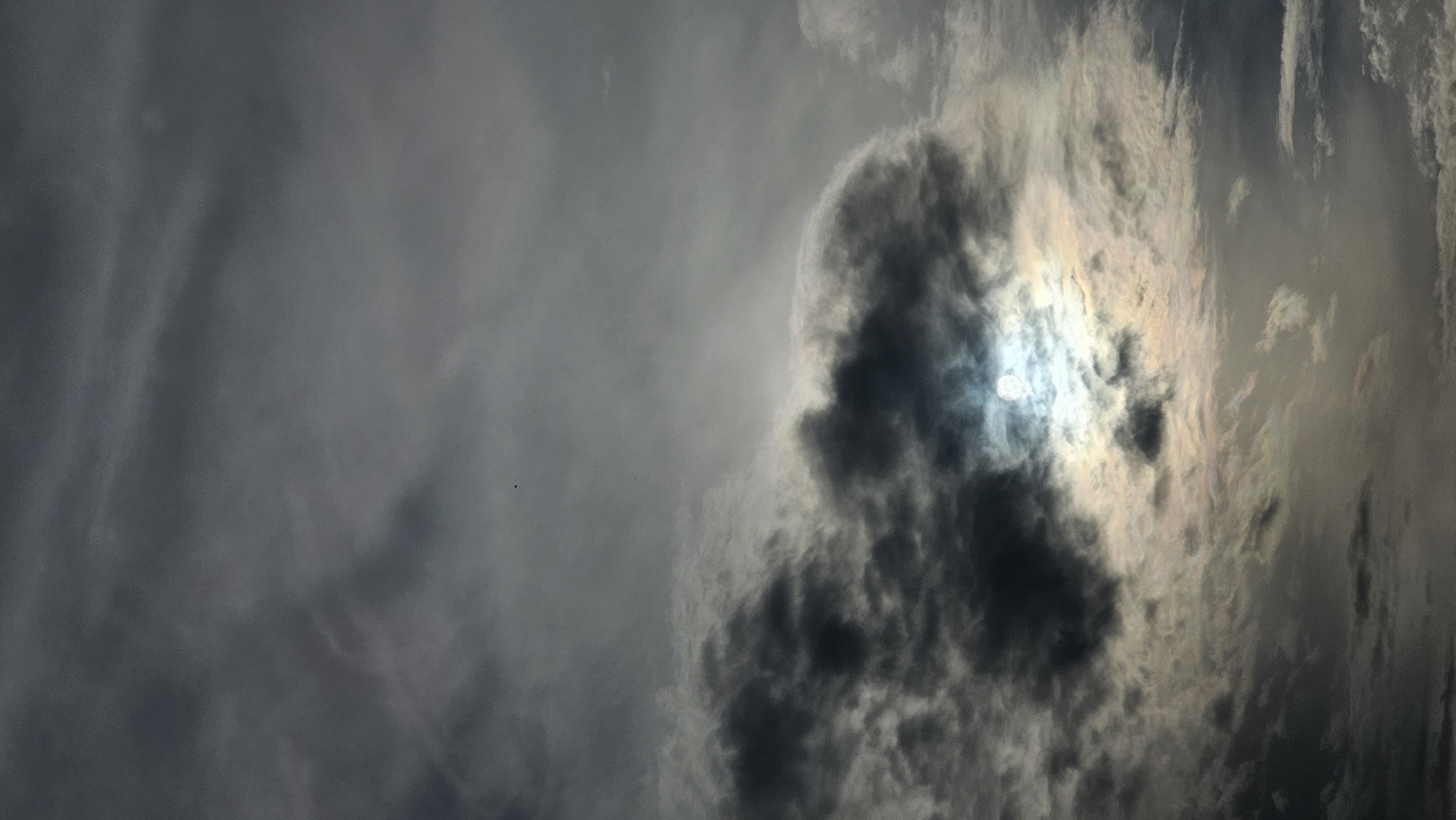 The image is a vertical, low-angle shot of a cloudy sky, captured from Kamensk-Uralsky, Russia. The sky is filled with dense, dark gray clouds, with some lighter, wispy clouds interspersed. The sun is partially obscured by a large, dark cloud mass in the lower center of the frame, creating a bright, hazy glow around its edges. The sun appears as a bright white disc with a halo effect due to the surrounding clouds. There are no visible people, animals, or man-made structures. The scene appears to be a natural outdoor environment, likely during daytime. A very small dark speck, possibly a bird or an insect, is visible in the upper portion of the sky.
