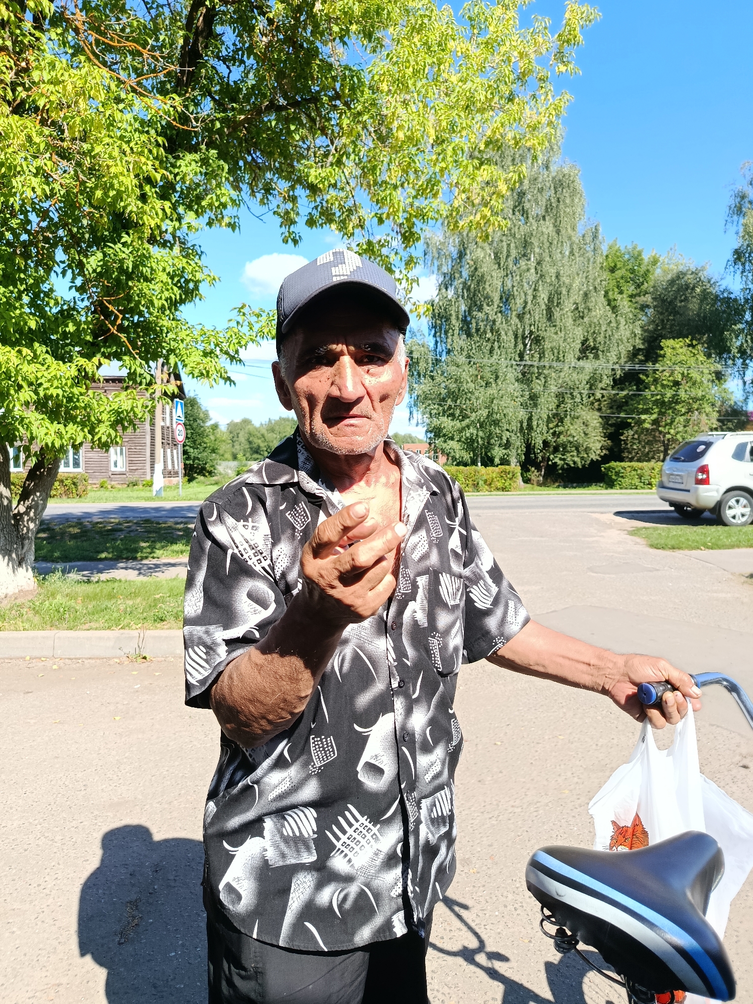 An older man with tanned skin and grey hair stands outdoors, holding a bicycle with his right hand, and a white plastic bag in his left. He is wearing a black and white patterned short-sleeved shirt and a dark baseball cap. His left hand is extended towards the camera, holding a small, light-colored object, possibly a piece of food. The man is looking directly at the camera with a serious expression. The background shows a street with a sidewalk and a grassy area. A wooden house and a street sign are visible to the left, and trees and a silver car are in the distance to the right. The sky is clear and blue. The scene appears to be set in the city of Vereya, Russia, given the context of the location.