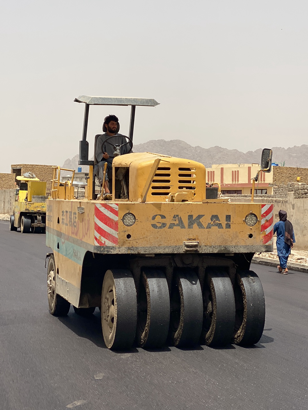 The image depicts a road construction scene in Kandahar, Afghanistan. A man with a beard is operating a yellow Sakai brand road roller on a newly paved asphalt road. The roller has multiple black tires for compacting the surface. In the background, another similar yellow roller is visible, along with buildings and mountains. A woman wearing a blue dress and headscarf is walking along the edge of the road.