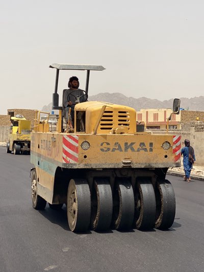 The image depicts a road construction scene in Kandahar, Afghanistan. A man with a beard is operating a yellow Sakai brand road roller on a newly paved asphalt road. The roller has multiple black tires for compacting the surface. In the background, another similar yellow roller is visible, along with buildings and mountains. A woman wearing a blue dress and headscarf is walking along the edge of the road.