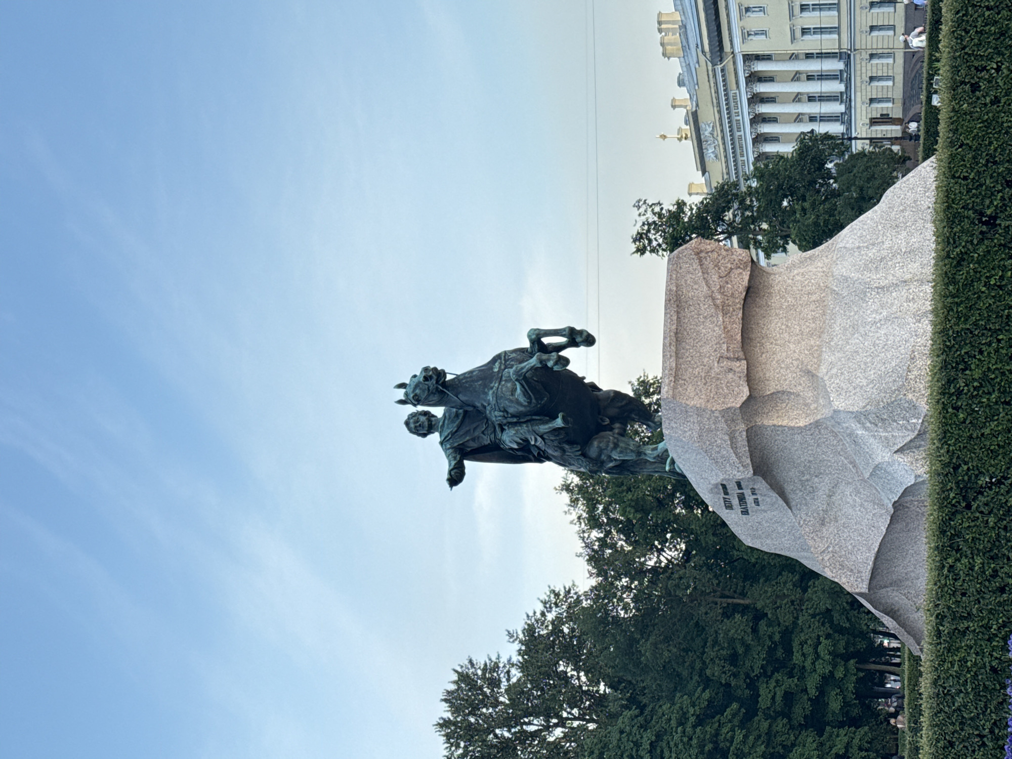 The image shows the Bronze Horseman, a statue of Peter the Great on horseback, located in Senate Square in Saint Petersburg, Russia. The equestrian statue, made of bronze, is mounted on a large granite base. Peter the Great is depicted with his right arm extended, pointing towards the Neva River. The statue is surrounded by trees and a manicured hedge. In the background, a neoclassical building with columns and a yellow facade is visible, likely the Senate and Synod buildings. The sky is light blue with scattered clouds. A few people are present in the background, appearing as small figures in the distance. The overall scene suggests an outdoor public space in an urban environment.