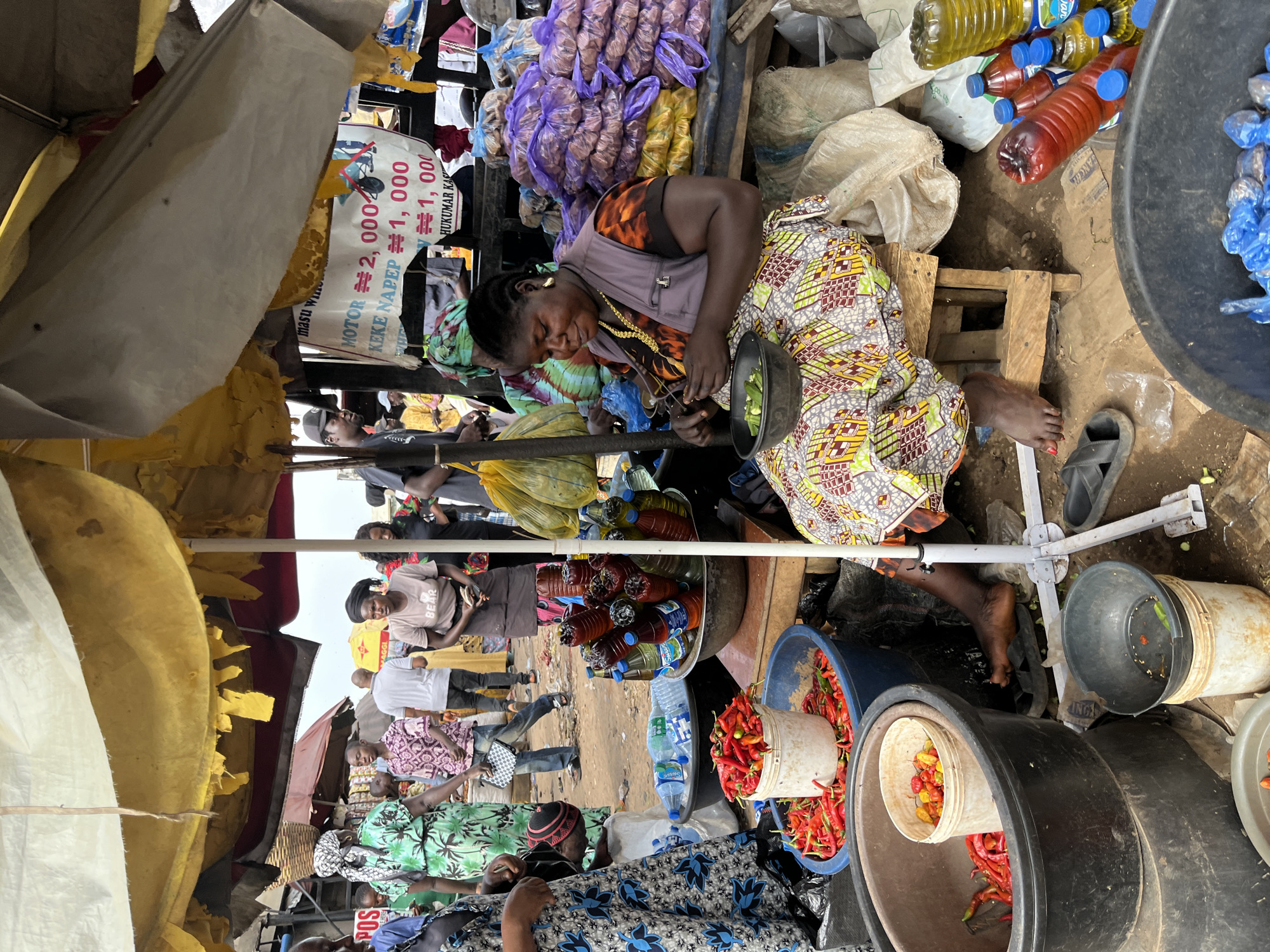 The image captures a vibrant scene at an outdoor market in Rafin Par, Nigeria. In the foreground, a woman is seated on a wooden stool, meticulously preparing food, likely okra, as evidenced by the bowl of green pods in her lap. She is wearing a colorful, patterned dress and a dark vest. To her right, several bottles of oil and water are neatly arranged, alongside large sacks of produce, possibly onions, wrapped in purple plastic. A variety of chilies, both red and yellow, are prominently displayed in large plastic tubs and buckets, indicating their availability for sale.
In the background, other market-goers are visible, engaged in their daily activities. A woman in a green patterned dress, possibly a vendor, stands to the left, while others are seen browsing or moving through the bustling marketplace. The market is shaded by a large yellow tarp, offering respite from the sun. A sign advertising prices for "MOTOR" and "KEKE NAPEP" is also visible, suggesting the presence of transportation services for hire. The ground is dirt and scattered with debris, typical of an open-air market environment. The overall atmosphere is one of lively commerce and everyday life in a Nigerian town.