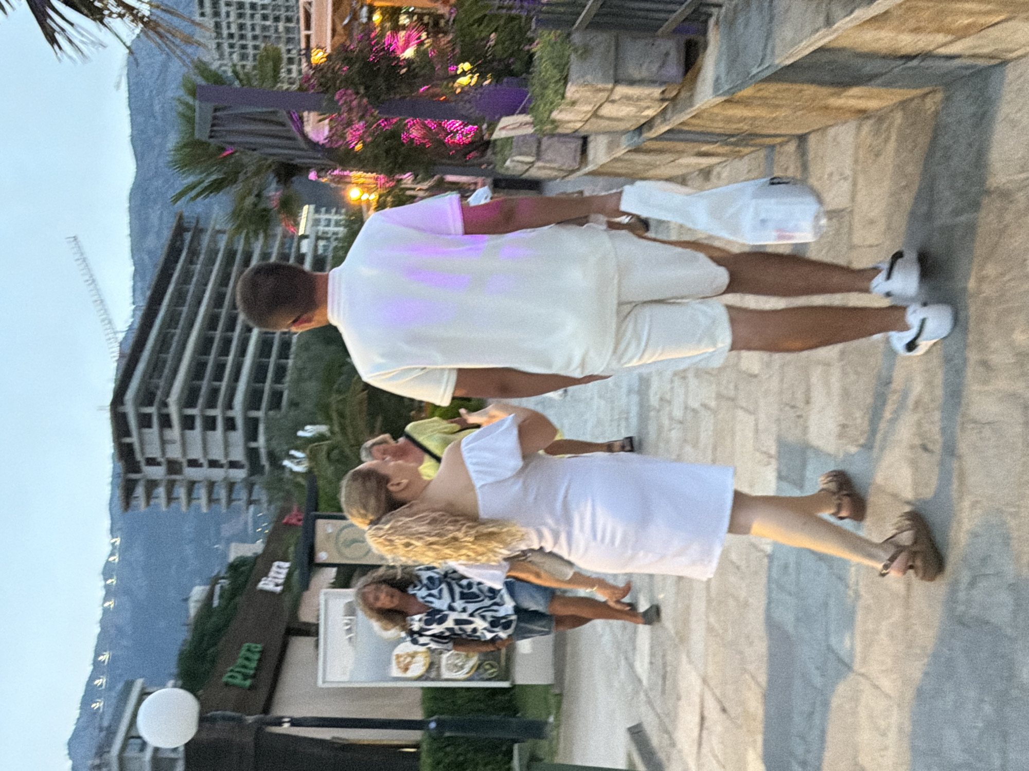 The image shows a street scene in Budva, Montenegro, at dusk. In the foreground, a couple walks away from the camera. The man is wearing a white polo shirt and white shorts, carrying a white plastic bag in his right hand. The woman is wearing a white off-the-shoulder dress and brown sandals. Behind them, a woman with blonde curly hair is walking towards the camera, wearing a patterned shirt and denim shorts. Another woman in a yellow top is further in the background. To the left of the frame, a building with a "Pizza" sign is visible. In the background, there is a multi-story building under construction and a mountainous landscape. The lighting suggests it is twilight, with artificial lights illuminating parts of the scene.