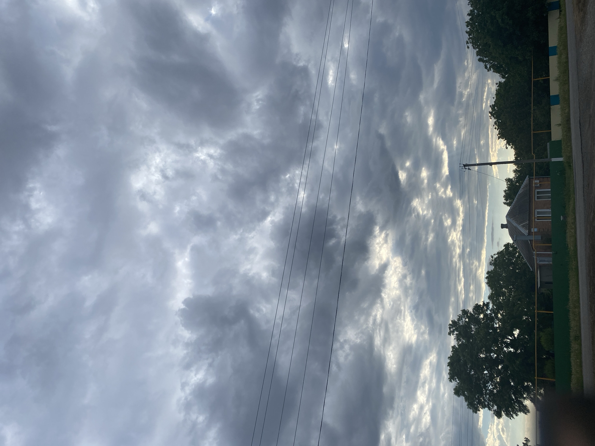 The image captures a landscape scene dominated by a dramatic, cloudy sky. The clouds are a mix of dark, brooding grays and lighter, brighter patches where the sun attempts to break through, creating a dynamic interplay of light and shadow. Several dark power lines stretch diagonally across the frame, cutting through the sky and adding a man-made element to the natural scene.
In the lower right portion of the image, a portion of a building with a tiled roof and brick walls is visible, suggesting a rural or suburban setting. Adjacent to the building are trees with dark green foliage. A fence, partially visible, with yellow and blue markings separates the property from a road. The lighting suggests it is either early morning or late afternoon, given the low angle of the sun implied by the bright patches in the clouds and the overall mood. The weather appears to be overcast, with a potential for rain or recent rain, given the dark clouds. There is no text visible in the image. The scene evokes a sense of quiet, possibly a slightly melancholic or contemplative atmosphere due to the heavy cloud cover.
